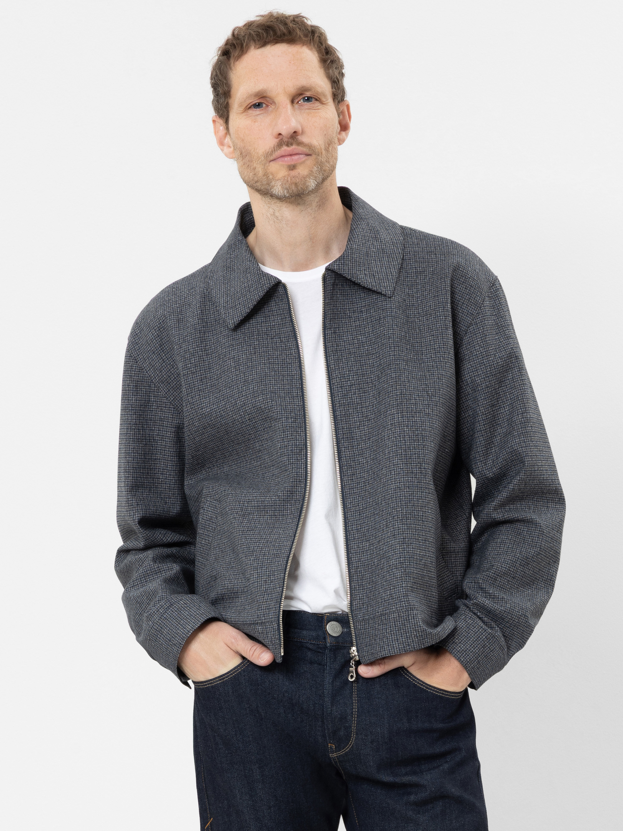 Ein Mann mit kurzen hellbraunen Haaren, der eine graue Jacke mit Reißverschluss über einem weißen T-Shirt und dunkelblauen Jeans trägt, steht mit den Händen in den Taschen vor einem schlichten weißen Hintergrund.
