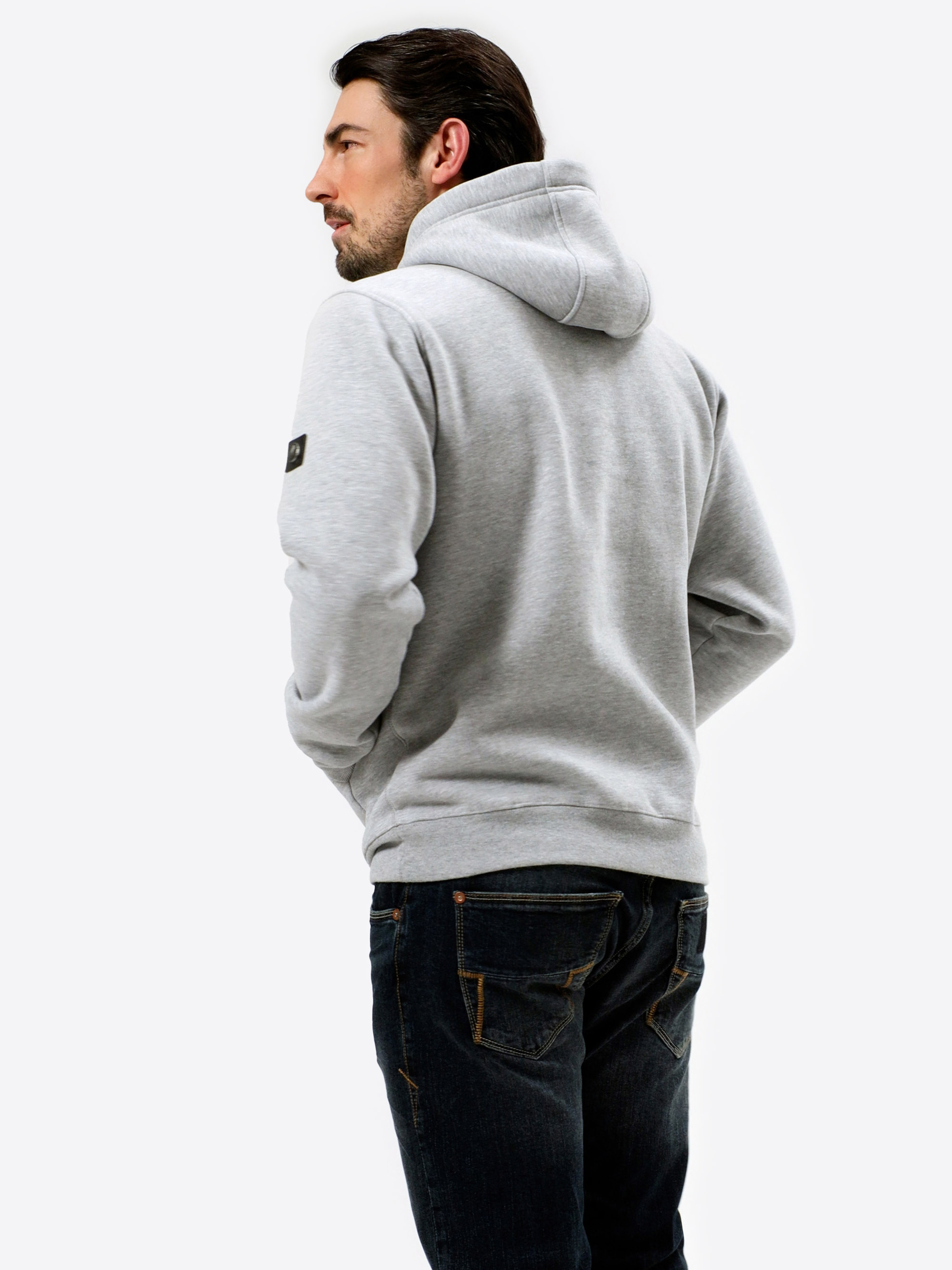 Ein Mann mit dunklem Haar, der ein hellgraues Kapuzensweatshirt und dunkelblaue Jeans trägt, steht mit dem Gesicht nach links vor einem schlichten weißen Hintergrund.