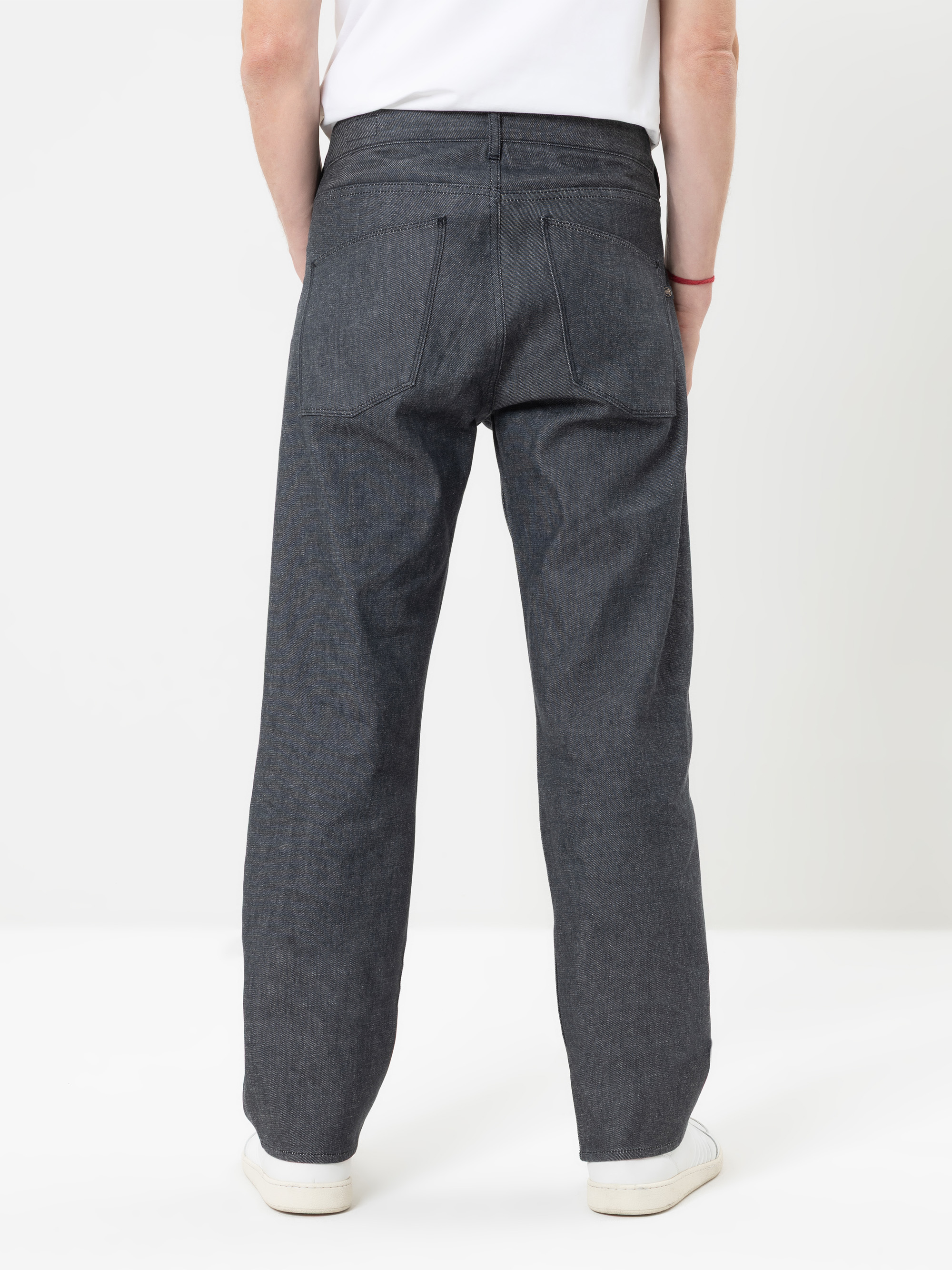 Eine Person, die eine dunkelgraue Jeans mit geradem Bein, weiße Turnschuhe und ein weißes Hemd trägt und mit dem Rücken zur Kamera vor einem schlichten weißen Hintergrund steht.
