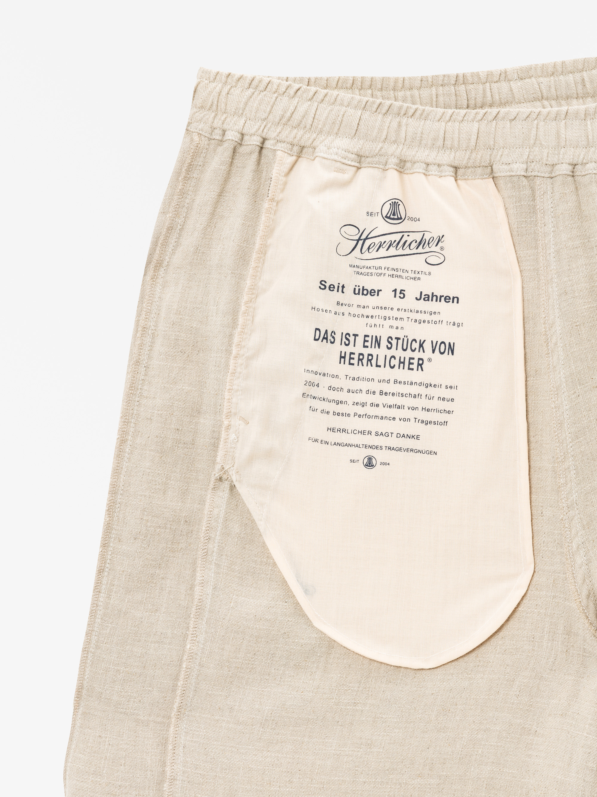 Nahaufnahme einer hellbeigen Hose mit einer Innentasche, die mit dem Markennamen Herrlicher und einem deutschen Text bedruckt ist, der die Qualität und die Geschichte der Marke beschreibt, und mit einem elastischen Bund am oberen Ende.