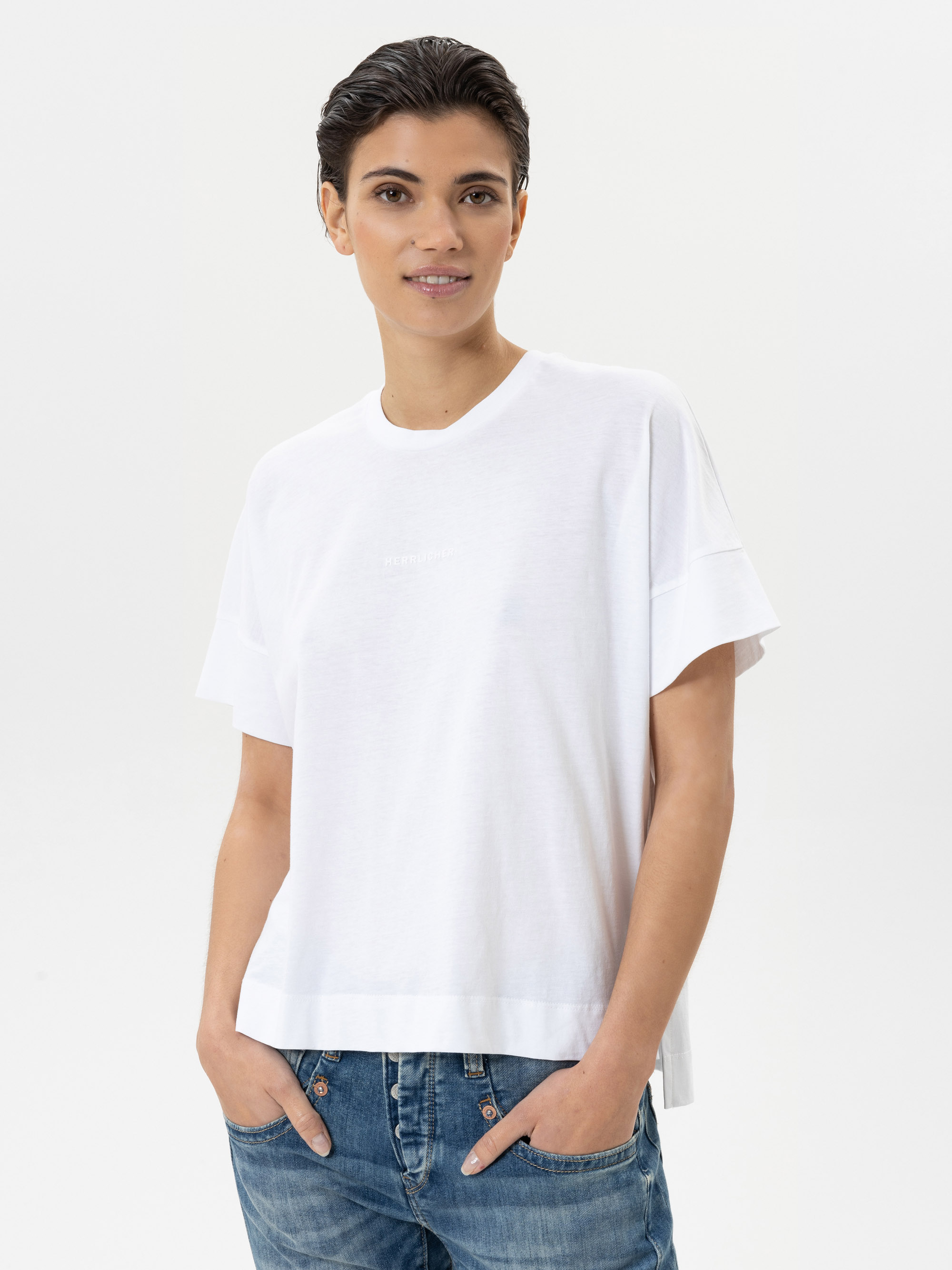 Eine Person mit kurzen dunklen Haaren, die ein lockeres weißes T-Shirt und blaue Jeans trägt, steht vor einem schlichten weißen Hintergrund, lächelt sanft und hat die Hände in den Taschen.