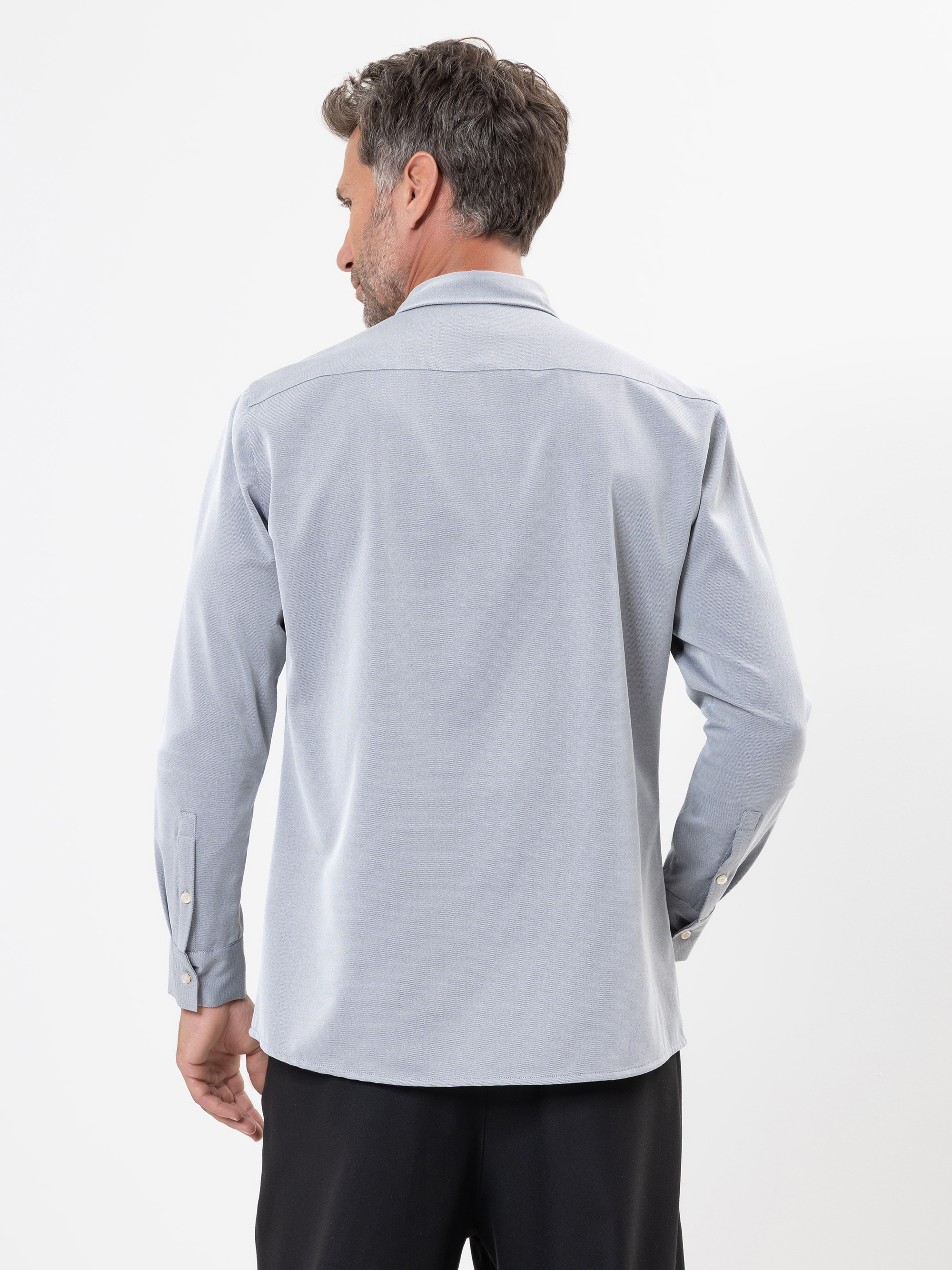 Ein Mann steht mit einem hellgrauen, langärmeligen Button-up-Hemd und einer schwarzen Hose vor einem einfarbig weißen Hintergrund und schaut von der Kamera weg.