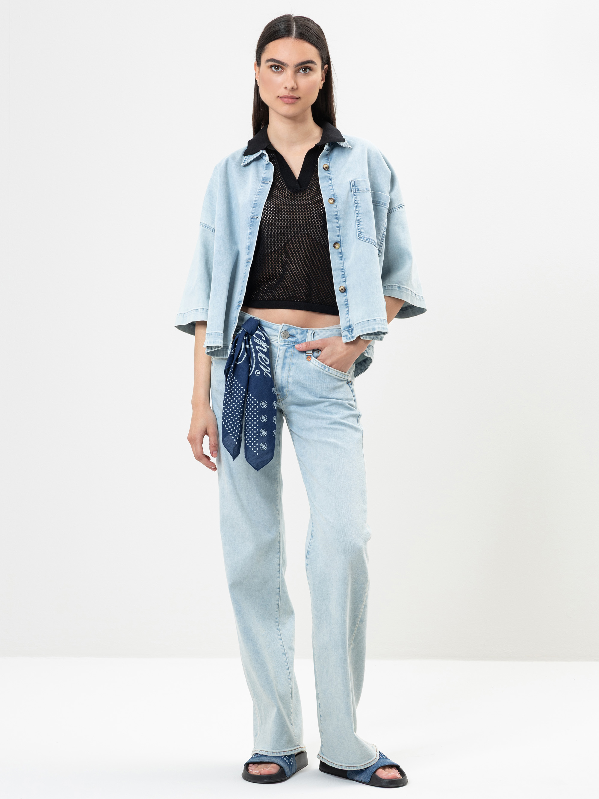 Eine Frau steht vor einem schlichten Hintergrund und trägt ein helles Jeanshemd und eine dazu passende Jeans, ein schwarzes Mesh-Top, dunkle Sandalen und ein blaues Halstuch, das sie sich an die Gürtelschlaufe gebunden hat. Sie hat langes dunkles Haar u