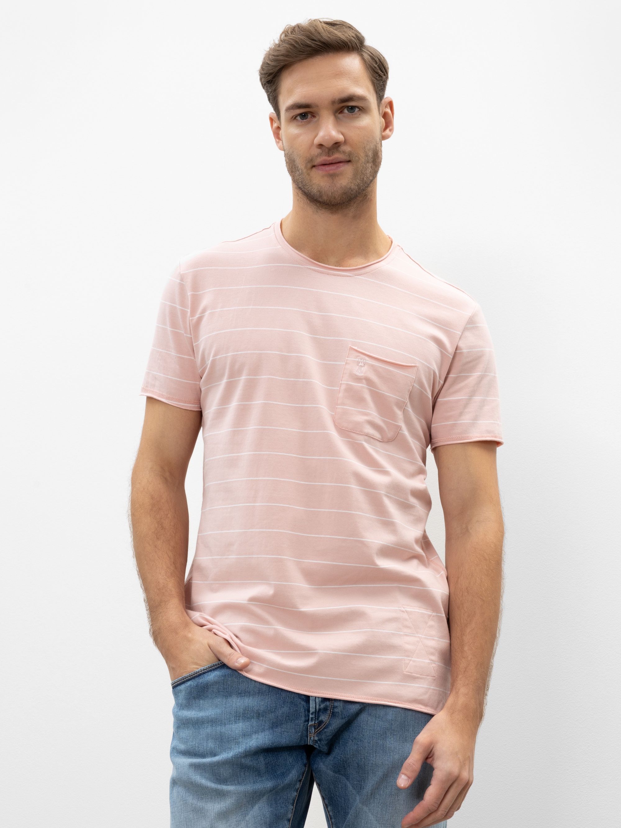 Ein Mann mit kurzen braunen Haaren und heller Gesichtsbehaarung trägt ein hellrosa, kurzärmeliges T-Shirt mit dezenten horizontalen Streifen und einer Brusttasche, gepaart mit einer blauen Jeans, vor einem schlichten weißen Hintergrund.