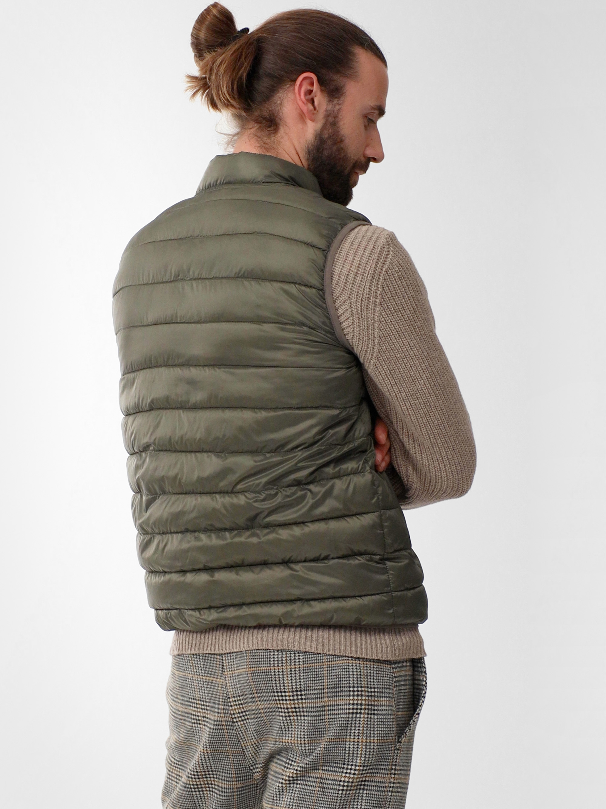 Ein Mann mit langen Haaren in einem Dutt trägt eine olivgrüne Pufferweste über einem beigen Pullover und einer karierten Hose und steht mit dem Rücken zur Kamera vor einem schlichten hellen Hintergrund.