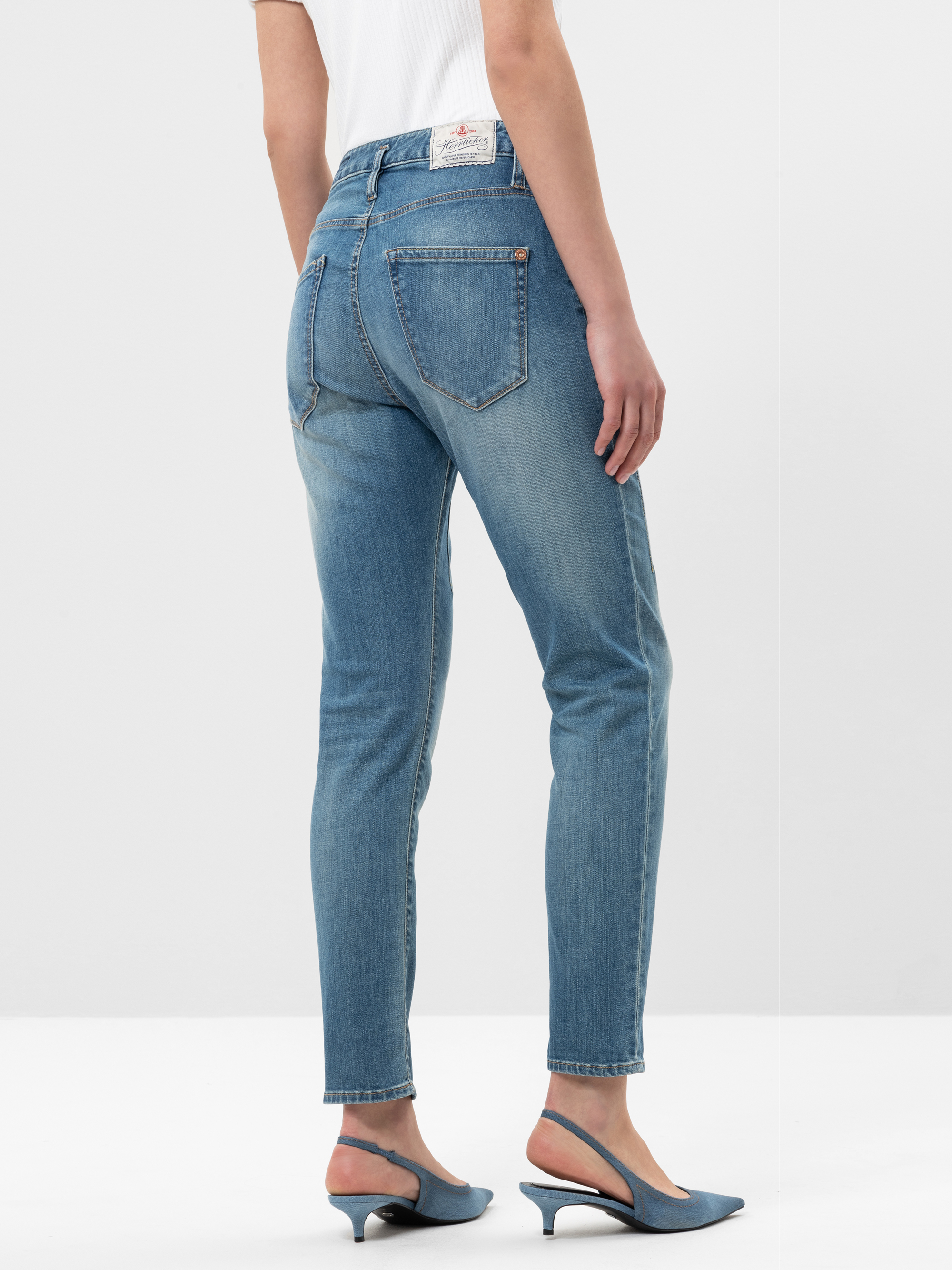 Eine Person, die hellblaue Jeans mit hoher Taille und blaue Slingback-Schuhe mit niedrigem Absatz trägt, steht mit dem Gesicht nach hinten und zeigt die Rückseite der Jeans und einen Teil eines weißen kurzärmeligen Oberteils vor einem unifarbenen Hint