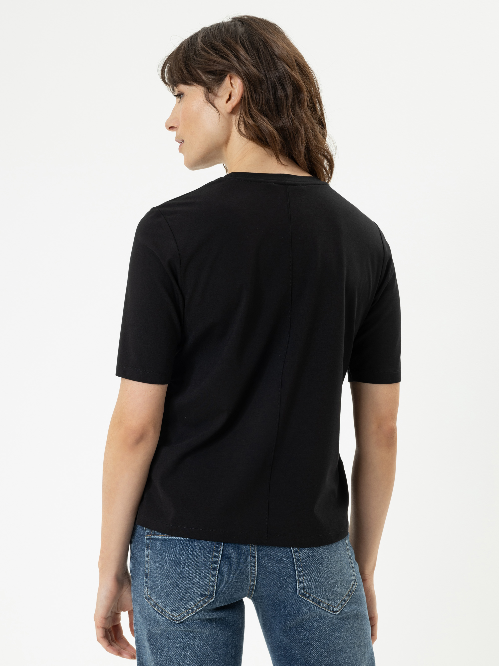 Eine Person mit gewelltem braunem Haar, die ein schwarzes kurzärmeliges T-Shirt und eine blaue Jeans trägt, steht mit dem Gesicht zur Seite vor einem schlichten weißen Hintergrund.