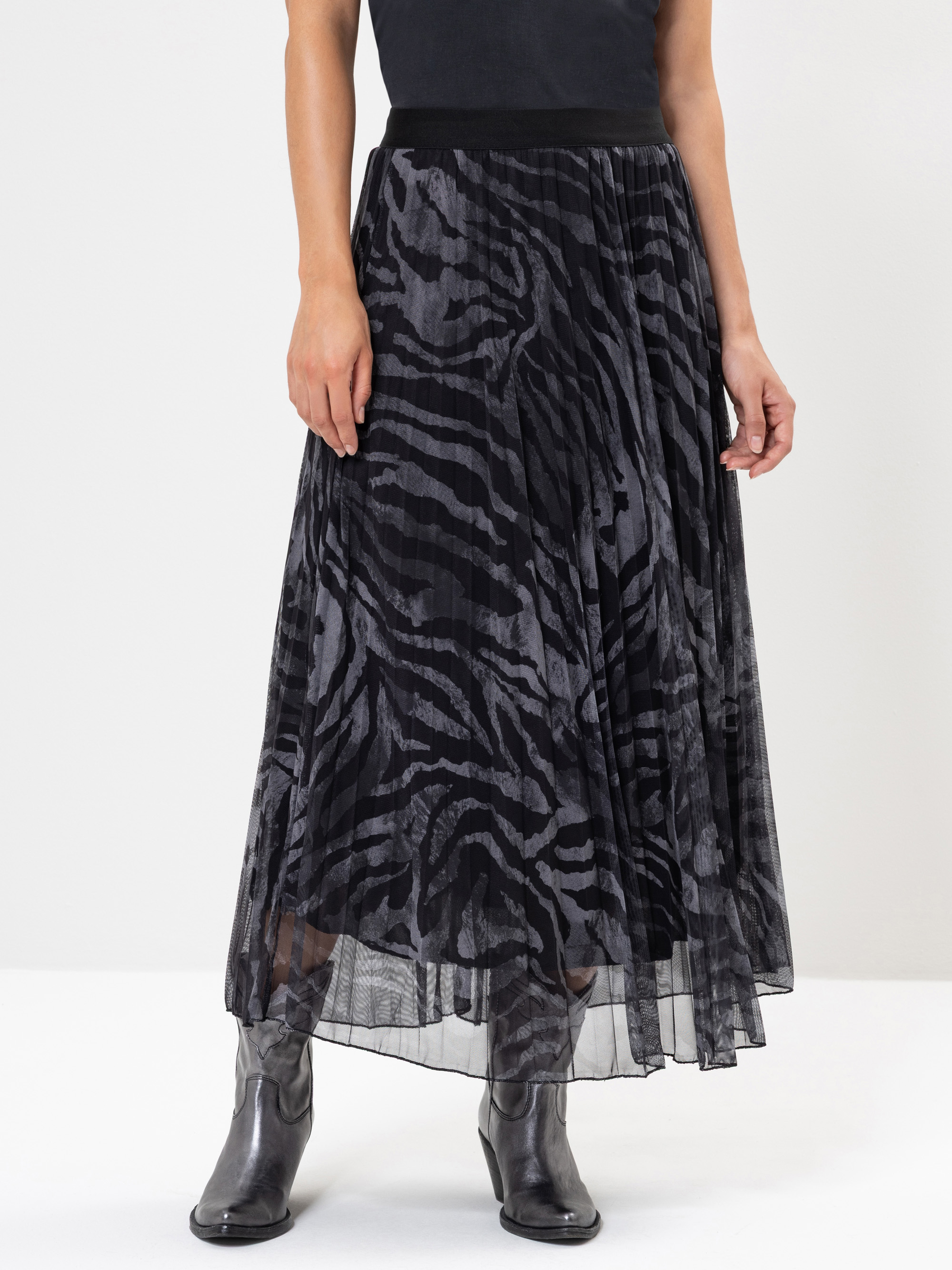 Eine Person, die ein schwarzes Oberteil, einen durchsichtigen Falten-Maxirock mit schwarzem und grauem Zebramuster und silberne Metallic-Stiefel trägt, steht vor einem schlichten hellen Hintergrund.