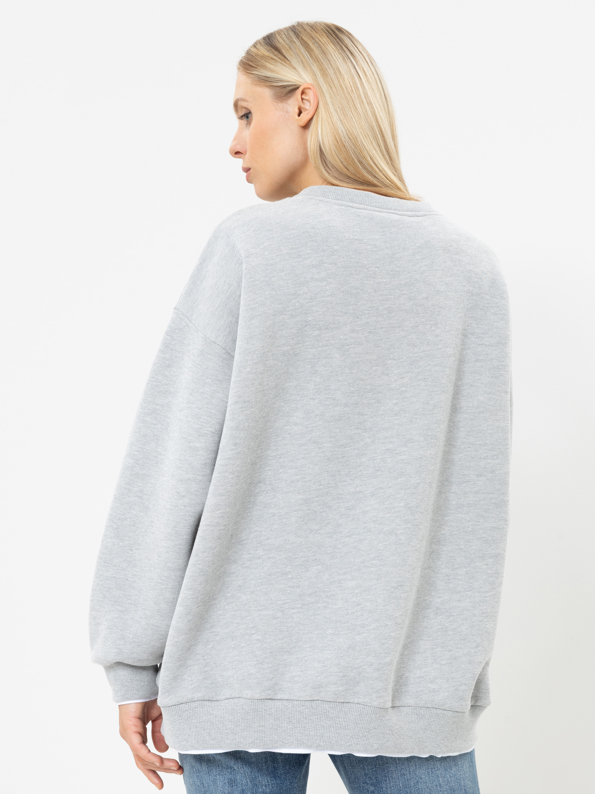 Eine Frau mit langen blonden Haaren trägt ein locker sitzendes hellgraues Sweatshirt und eine blaue Jeans und steht mit dem Rücken zur Kamera vor einem schlichten weißen Hintergrund.
