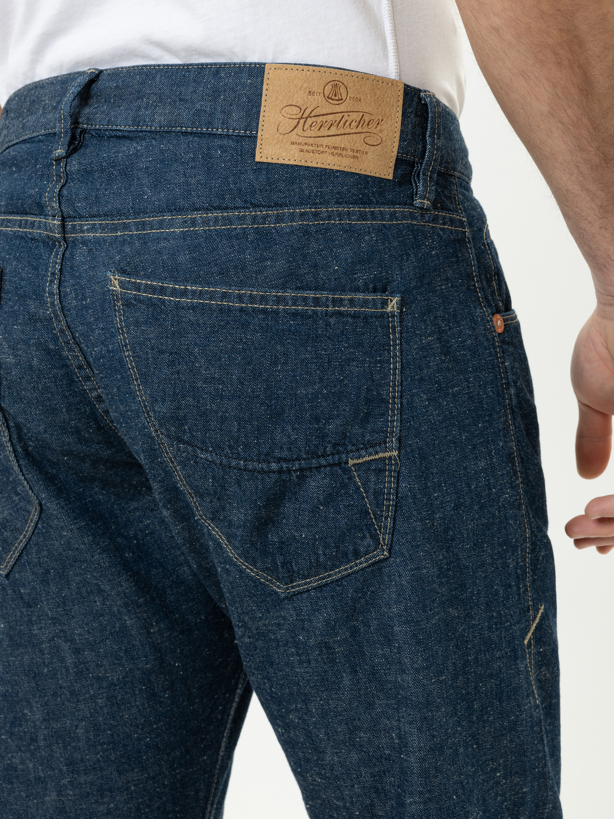 Nahaufnahme einer Person, die eine blaue Jeans mit hellbraunen Nähten und einem braunen Lederaufnäher am Bund mit der Aufschrift Herrlicher trägt. Nur der untere Rücken und die Oberschenkel sind sichtbar.