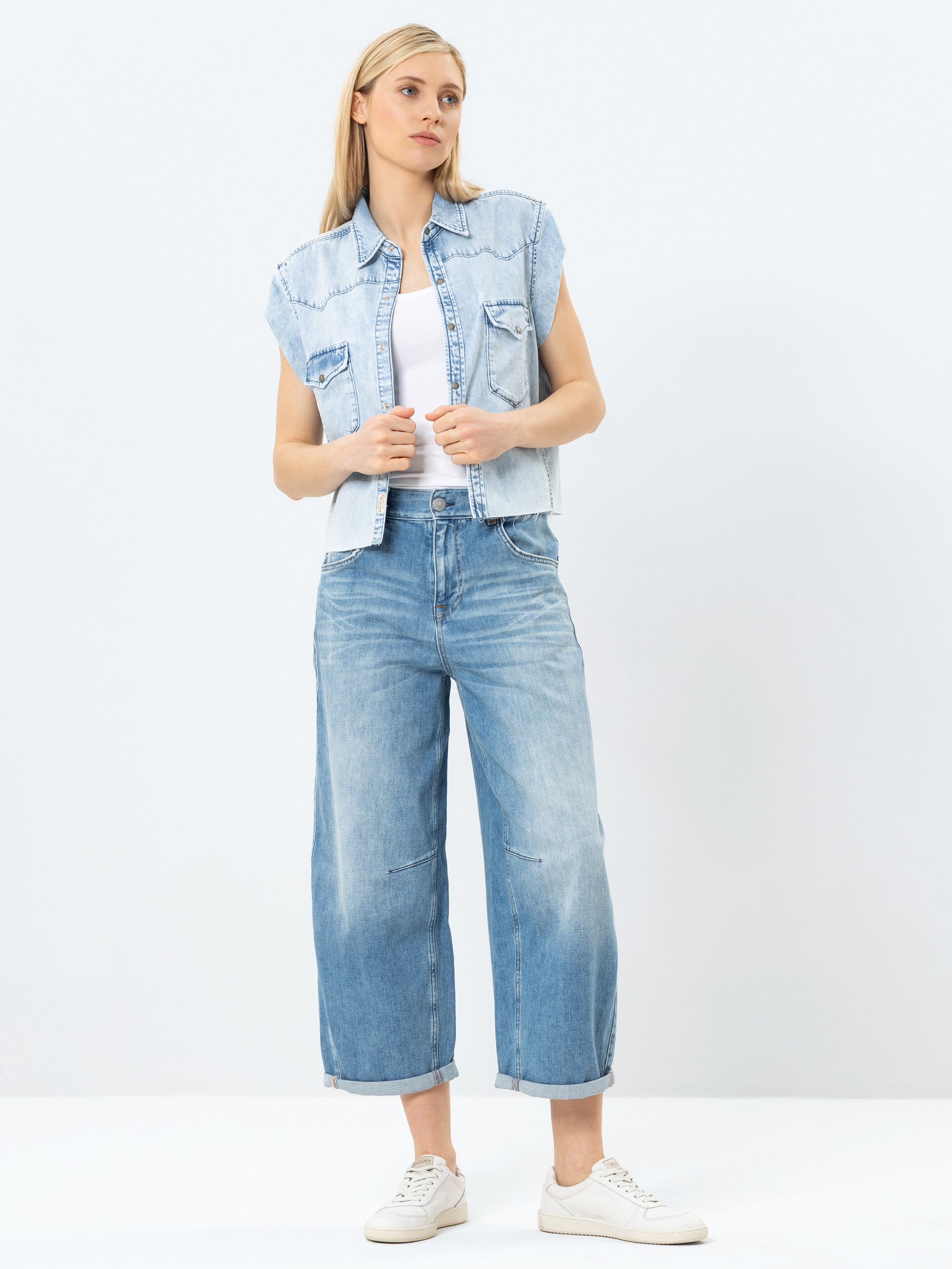 Eine Frau steht vor einem schlichten Hintergrund und trägt eine hellblaue Jeansweste über einem weißen Oberteil, Jeans mit weiten Beinbündchen und weiße Turnschuhe. Sie hat glattes, blondes Haar und einen neutralen Gesichtsausdruck.