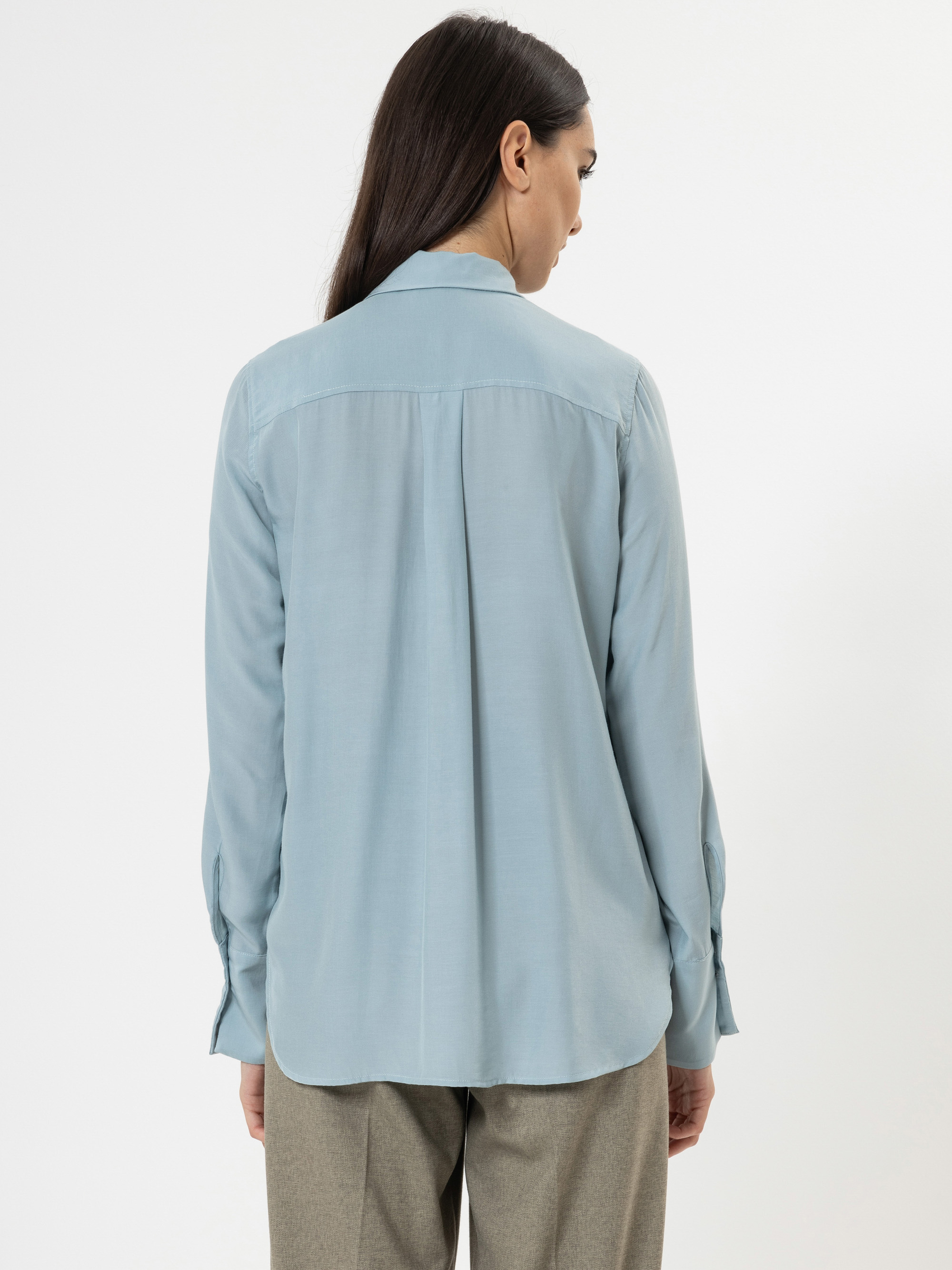 Eine Frau mit langen dunklen Haaren steht mit dem Rücken zur Kamera. Sie trägt ein hellblaues Button-up-Hemd und eine beigefarbene Hose vor einem schlichten weißen Hintergrund.