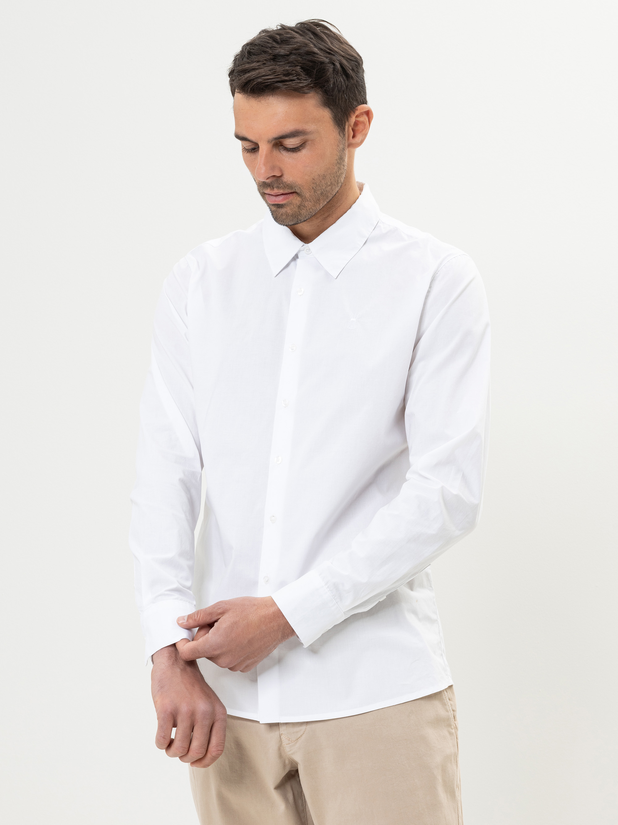 Ein Mann mit kurzen dunklen Haaren trägt ein weißes langärmeliges Hemd mit Knöpfen und eine beigefarbene Hose. Er blickt nach unten, während er seine linke Hemdmanschette vor einem schlichten hellen Hintergrund zurechtrückt.