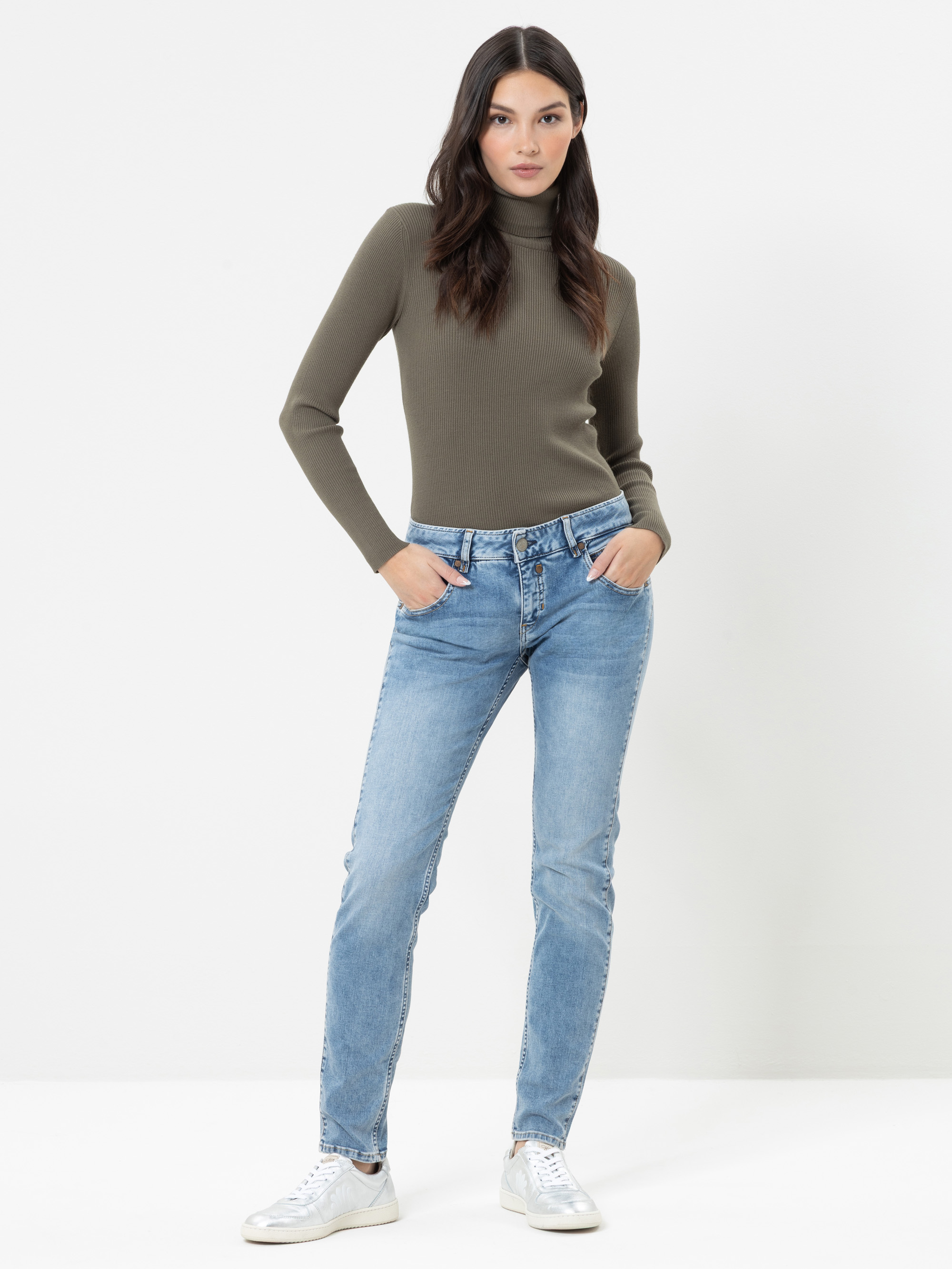 Eine Frau steht vor einem weißen Hintergrund und trägt einen eng anliegenden olivgrünen Rollkragenpullover, hellblaue Jeans und weiße Sneaker; sie hat die Hände in den Taschen und einen ausdruckslosen Gesichtsausdruck.