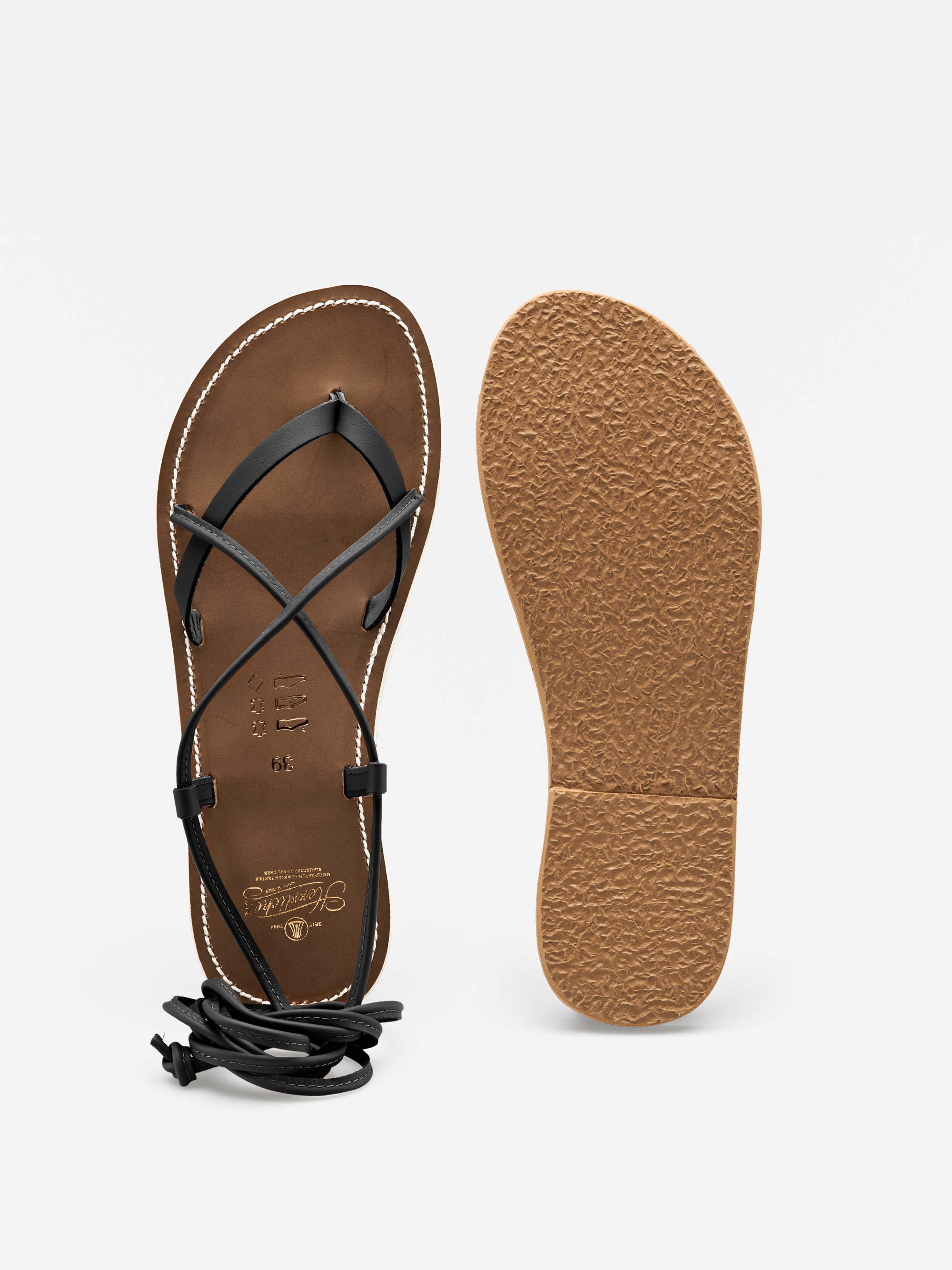 Ein Paar flache Sandalen mit brauner Sohle und schwarzen, gekreuzten Riemen. Eine Sandale zeigt die Draufsicht mit umgeschlagenen Knöchelriemen, während die andere die strukturierte Sohle zeigt.