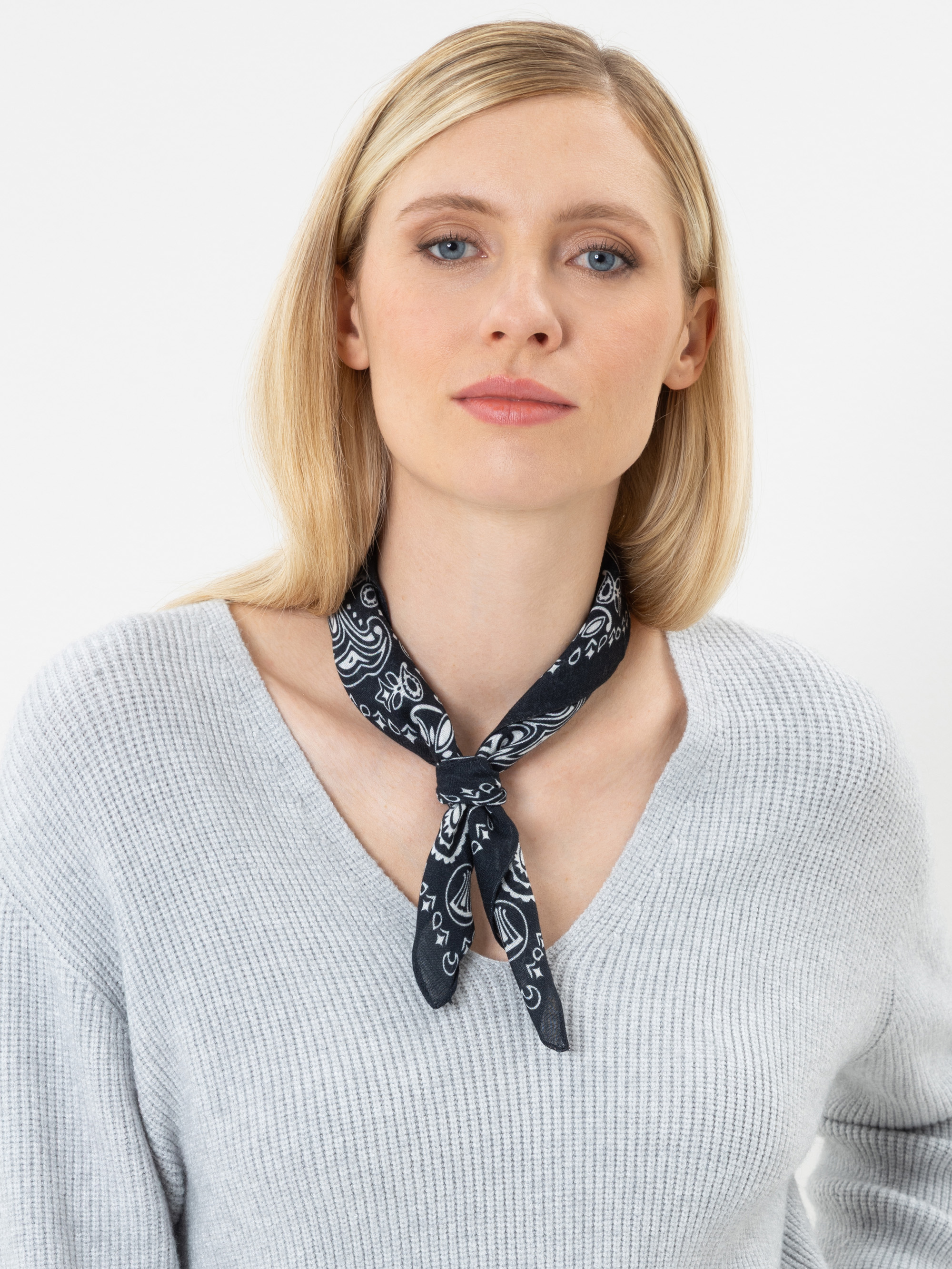 Eine Frau mit glattem blondem Haar trägt einen hellgrauen, gerippten Pullover und ein schwarz gemustertes Bandana, das sie sich um den Hals gebunden hat; sie steht vor einem schlichten weißen Hintergrund.