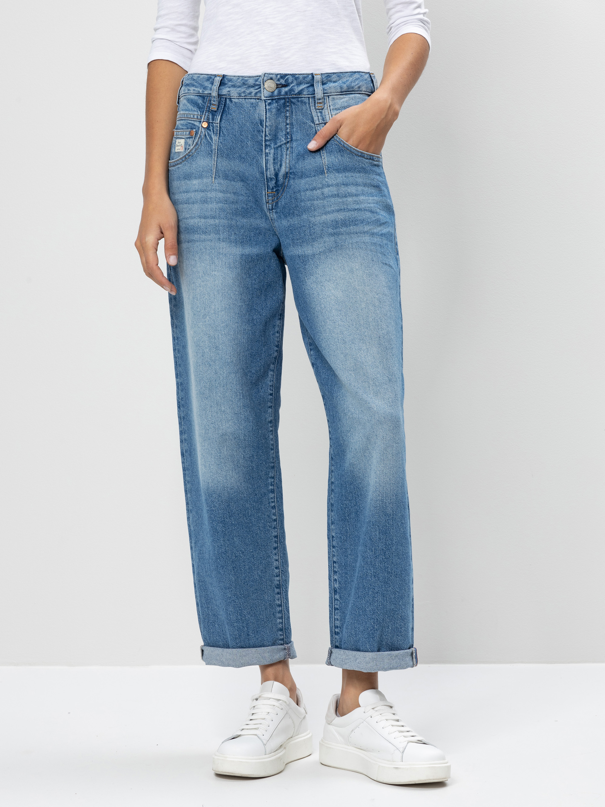 Eine Person, die eine hellblaue, locker sitzende Jeans mit aufgerollten Manschetten, ein weißes langärmeliges Oberteil und weiße Turnschuhe trägt, steht vor einem schlichten hellen Hintergrund. Ihre Hände befinden sich in den Taschen, und ihr Kopf is