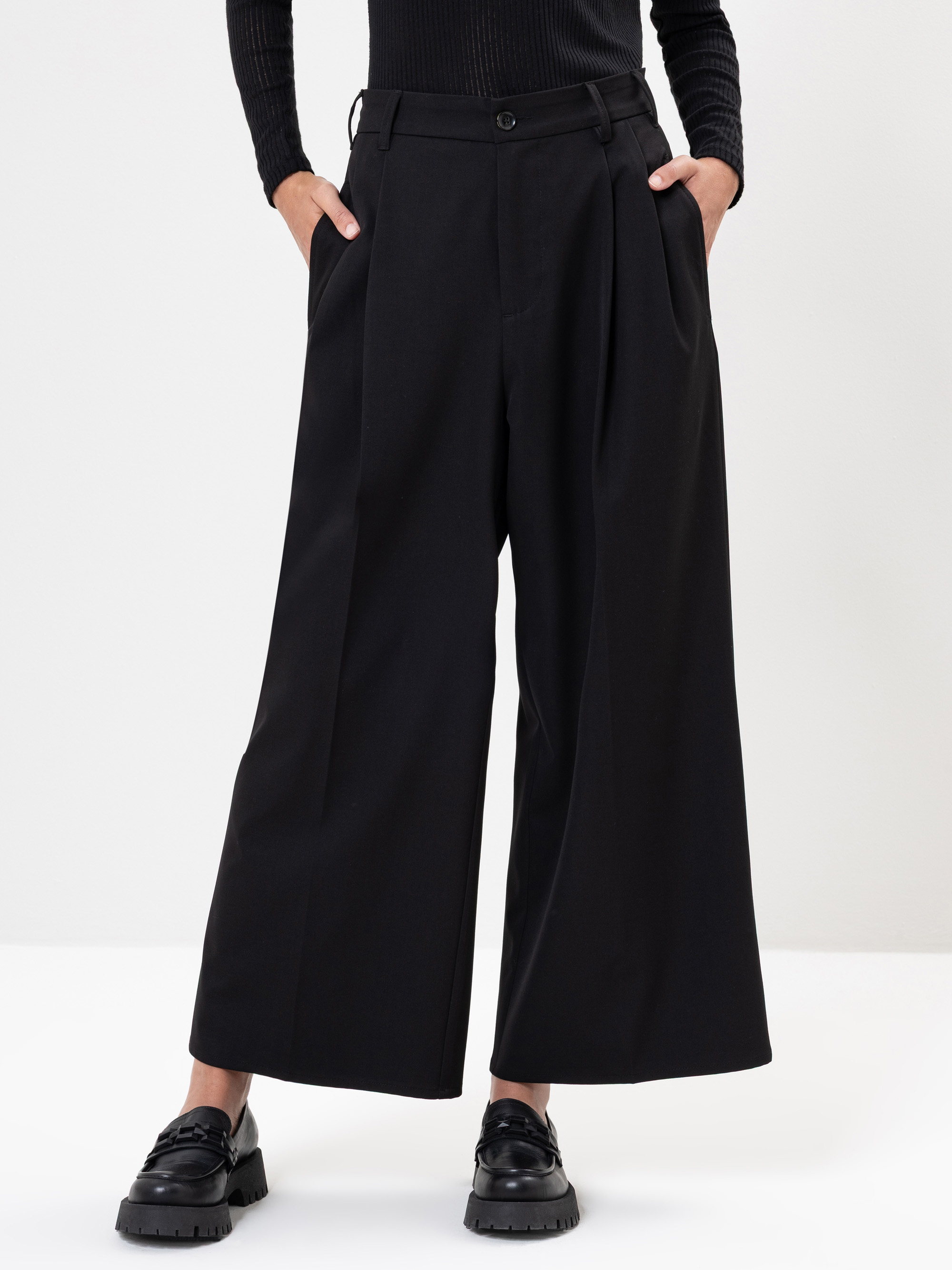 Person, die eine schwarze Hose mit weitem Bein, schwarze Halbschuhe und ein schwarzes langärmeliges Oberteil trägt, steht mit den Händen in den Taschen vor einem schlichten weißen Hintergrund.