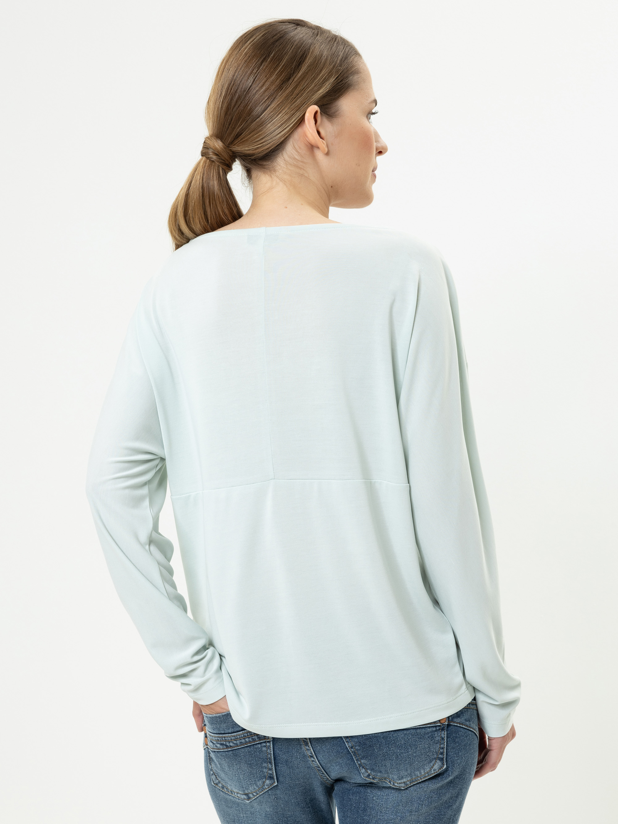 Eine Frau mit hellbraunem Haar in einem niedrigen Pferdeschwanz trägt ein hellblaues langärmeliges Oberteil und blaue Jeans und steht mit dem Rücken zur Kamera vor einem schlichten weißen Hintergrund.