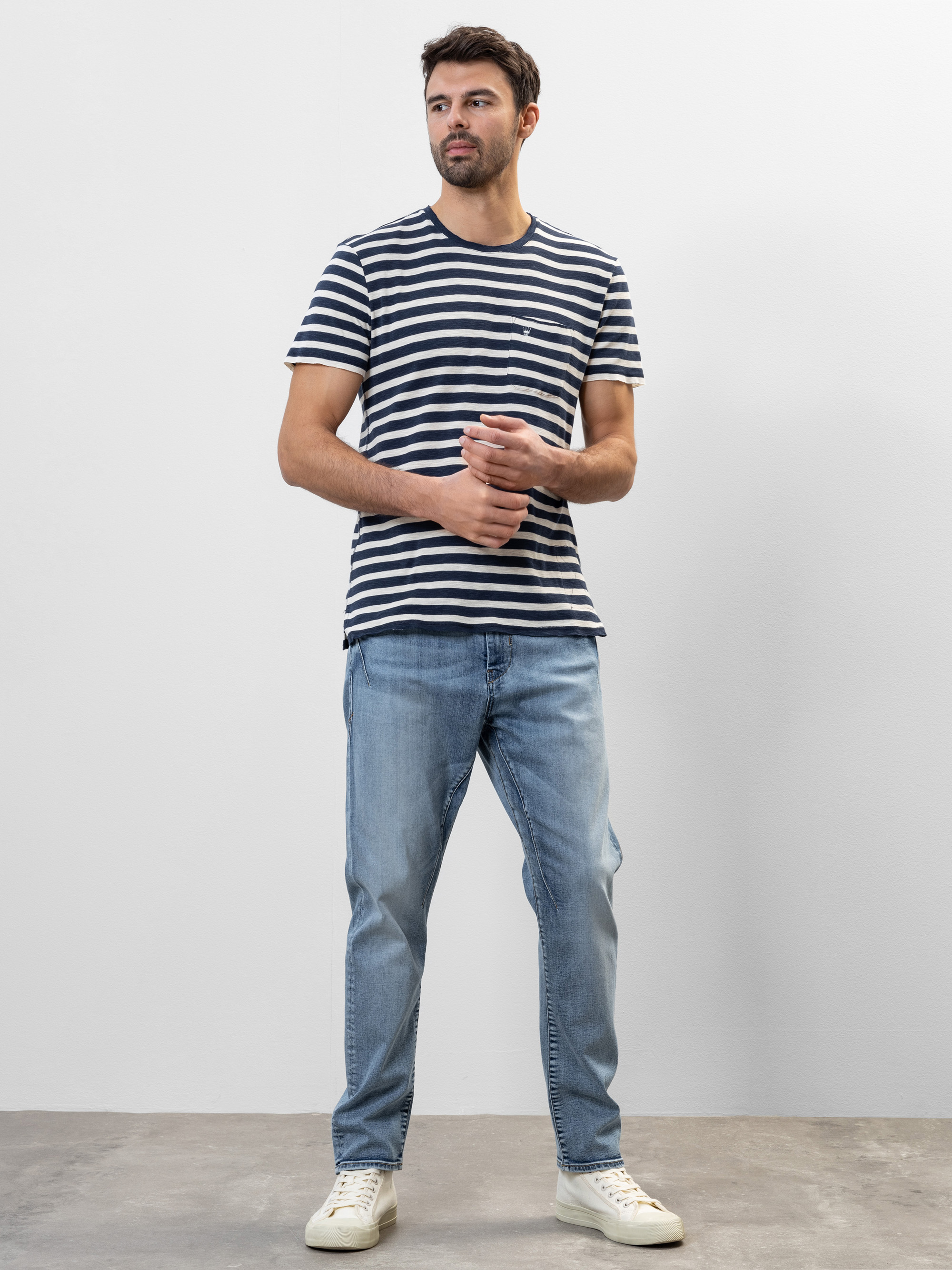 Ein Mann steht vor einer schlichten weißen Wand und trägt ein marineblau-weiß gestreiftes T-Shirt, hellblaue Jeans und weiße Turnschuhe. Er hat kurzes dunkles Haar und einen Bart und blickt mit locker gefalteten Händen leicht nach links.