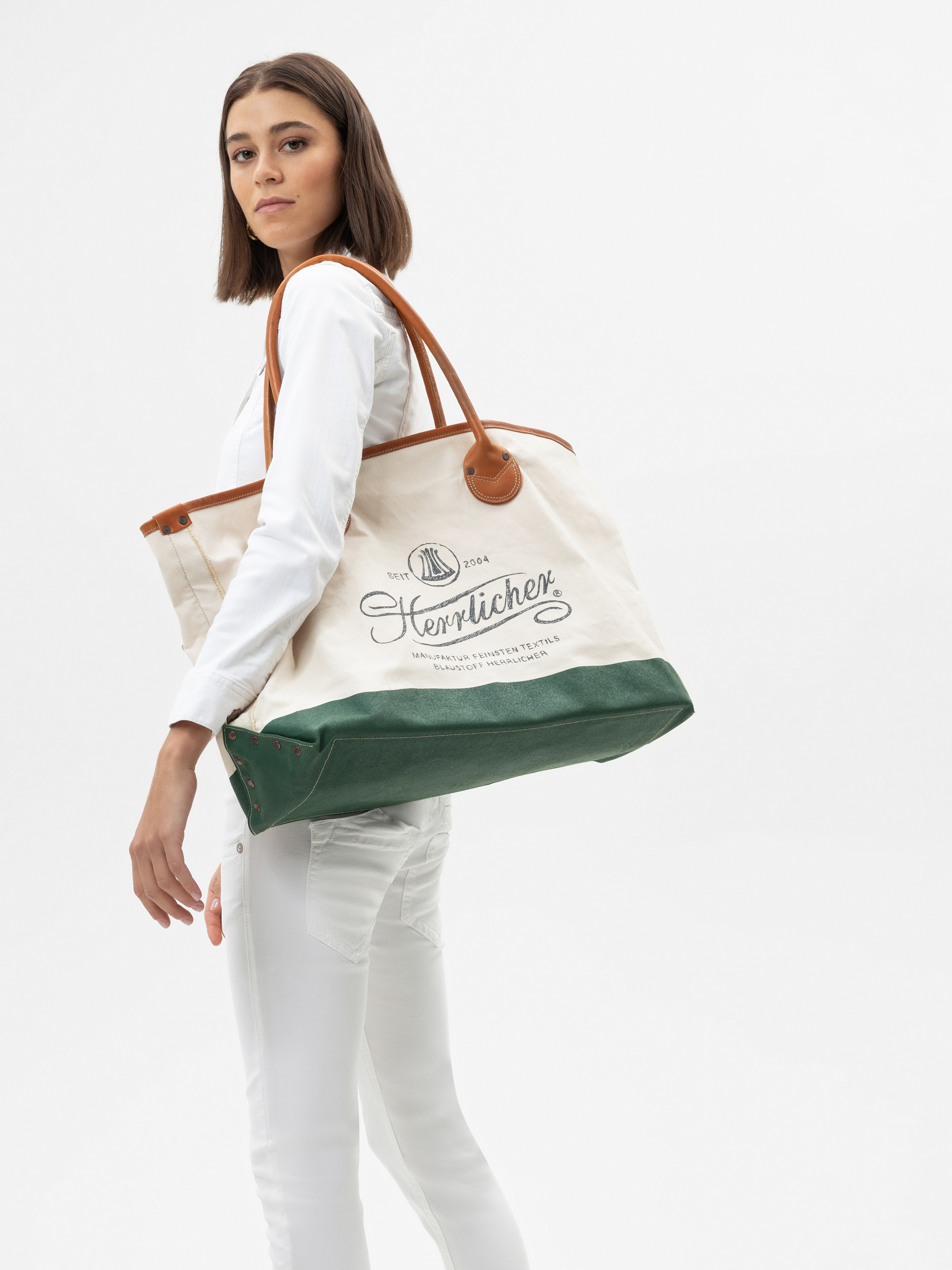 Eine ganz in Weiß gekleidete Frau hält eine große Tragetasche aus Segeltuch mit grünen Akzenten, braunen Ledergriffen und schwarzer Zierschrift auf der Vorderseite. Sie steht vor einem schlichten weißen Hintergrund und schaut über ihre Schulter.