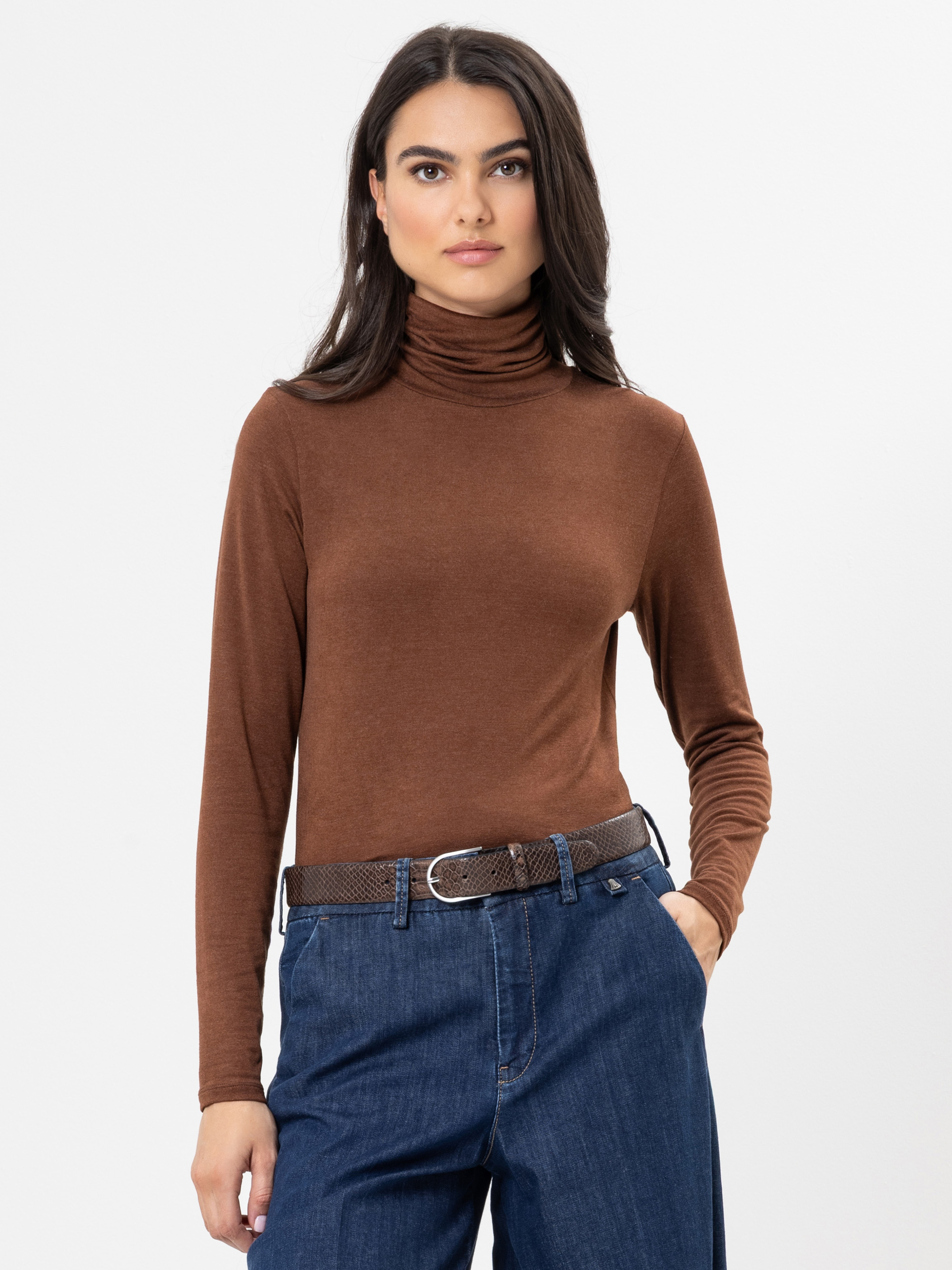 Eine Frau mit langen dunklen Haaren trägt einen braunen Rollkragenpullover, der in eine blaue Jeans mit hoher Taille und einem braunen Gürtel gesteckt ist, und steht vor einem schlichten weißen Hintergrund.