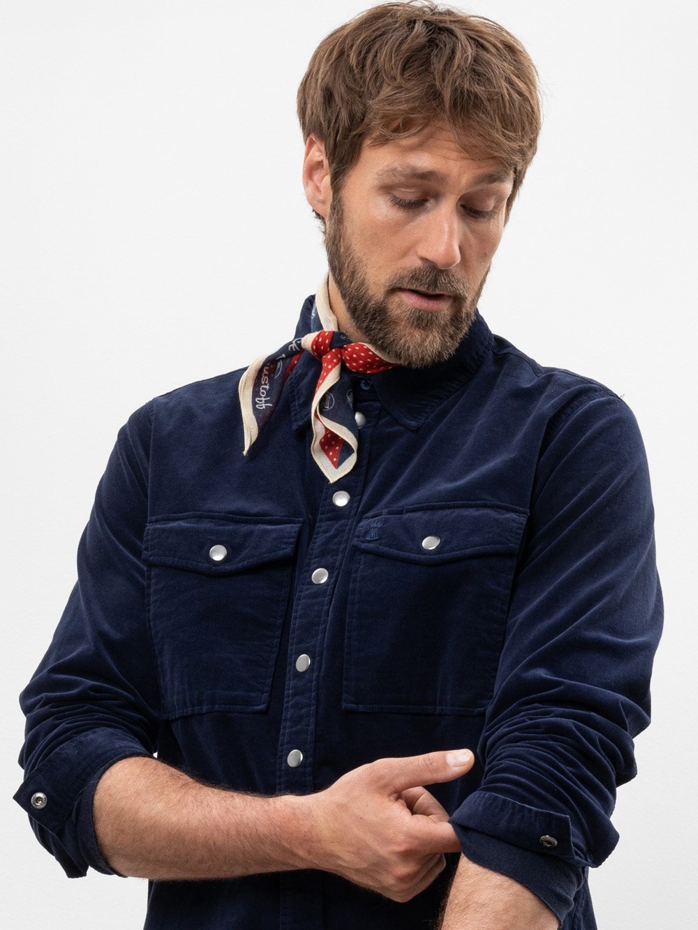 Ein Mann mit hellbraunem Haar und Bart krempelt den Ärmel seines marineblauen Button-up-Hemdes hoch. Er trägt einen gemusterten Schal um den Hals und blickt auf seinen Ärmel vor einem schlichten weißen Hintergrund hinunter.
