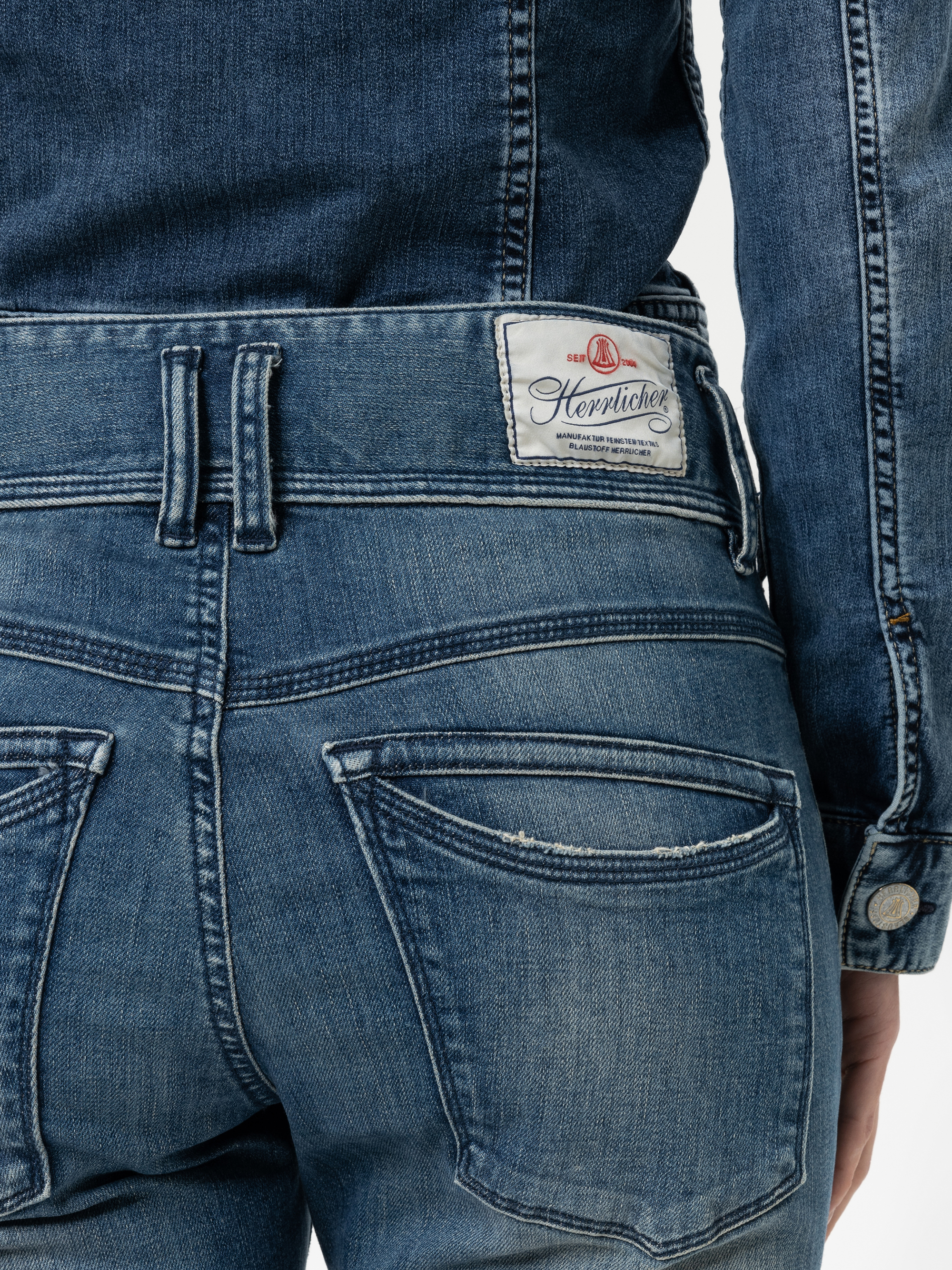 Nahaufnahme einer Person, die eine blaue Denim-Jeans und eine dazu passende Denim-Jacke trägt. Die Jeans hat ein weißes Herrlicher-Markenetikett über der rechten Gesäßtasche und sichtbare Nähte.