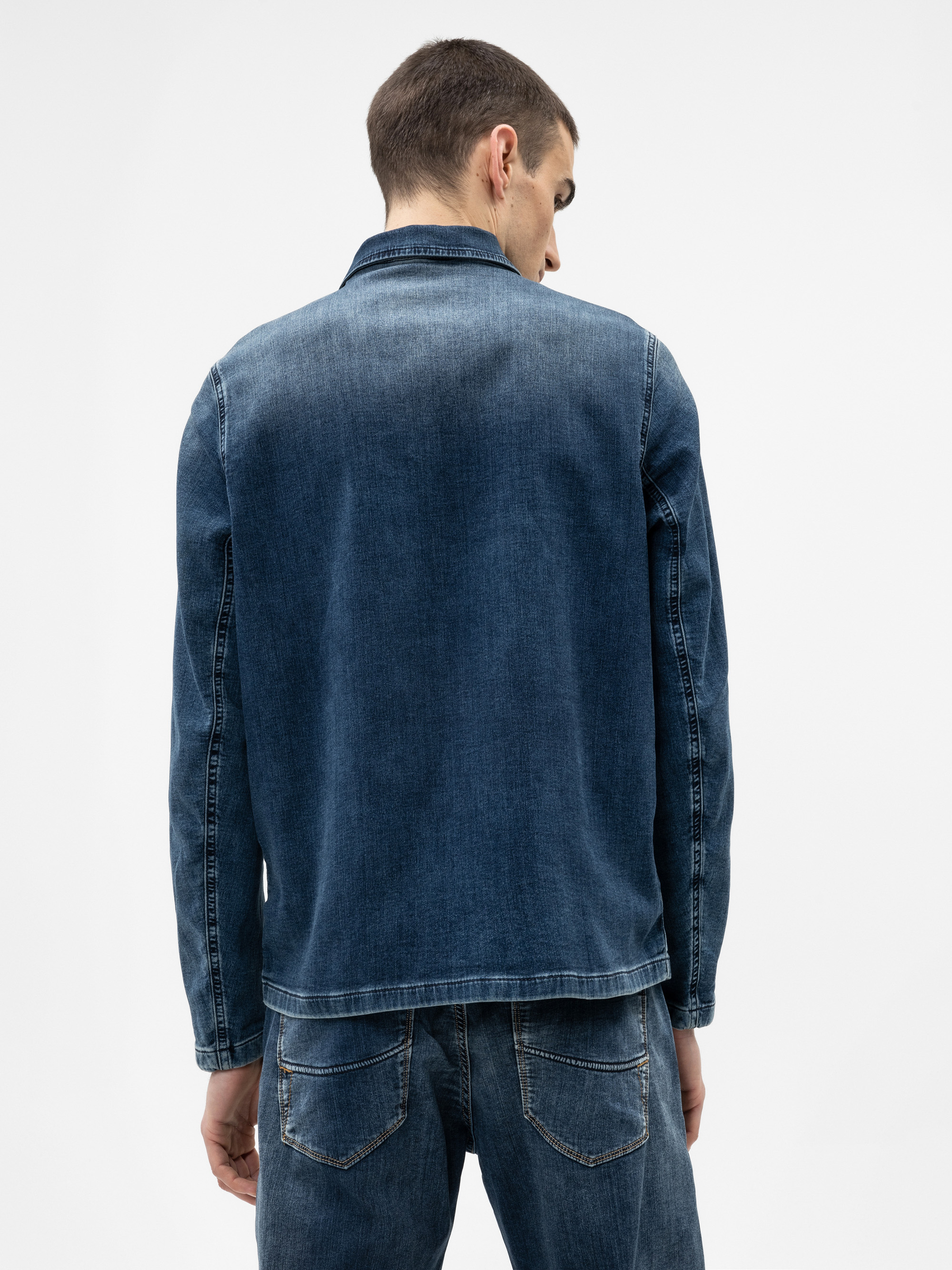 Ein Mann, der von der Kamera wegschaut, trägt eine blaue Jeansjacke und eine dazu passende blaue Jeans vor einem schlichten weißen Hintergrund.