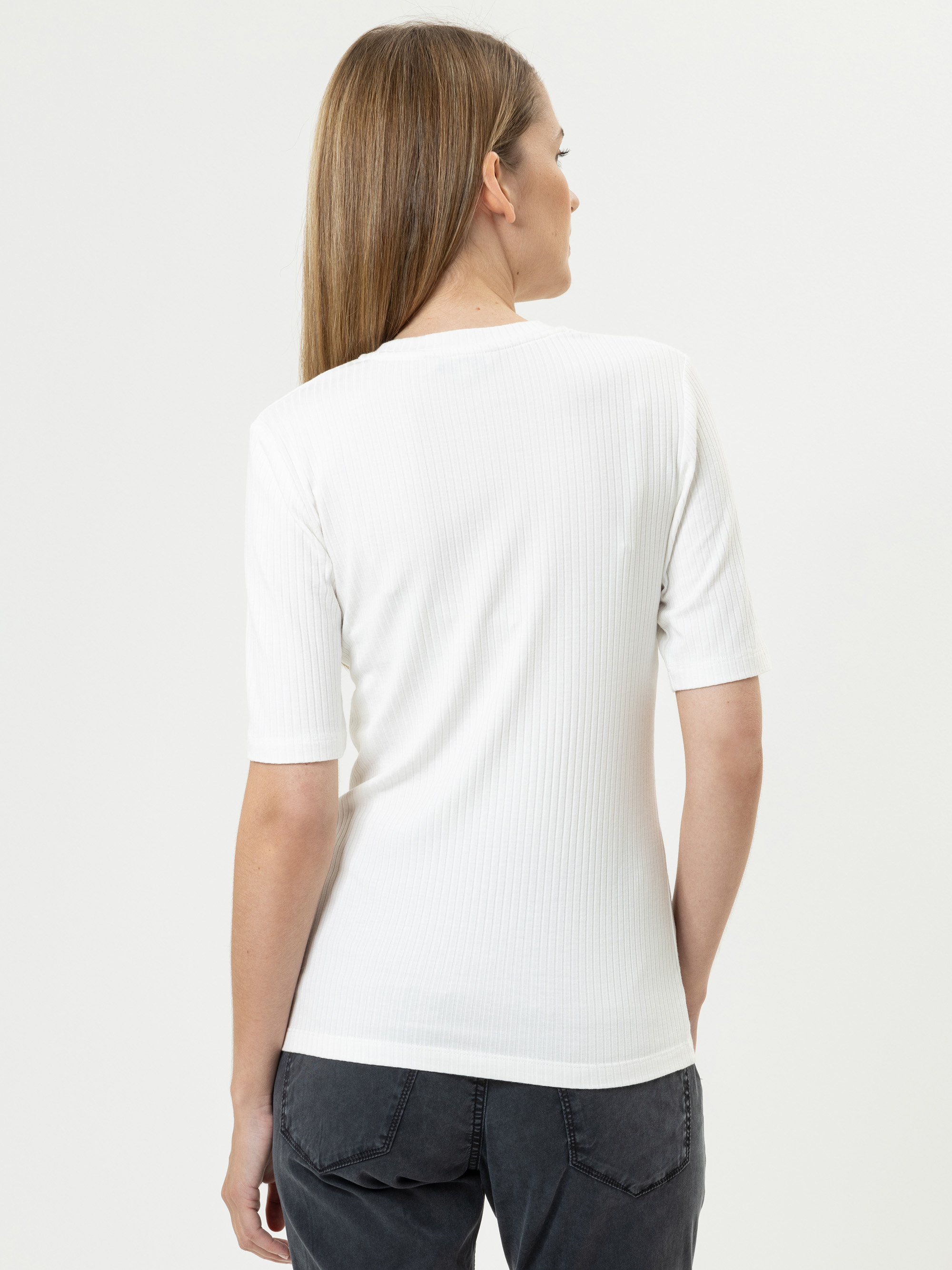 Eine Person mit glattem, hellbraunem Haar wird von hinten gezeigt. Sie trägt ein weißes, kurzärmeliges, geripptes Oberteil und schwarze Jeans vor einem schlichten weißen Hintergrund.