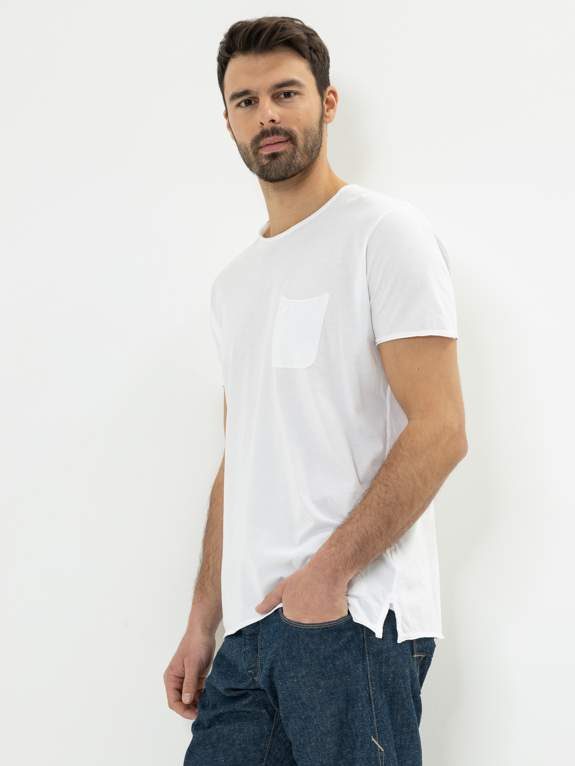Ein Mann mit dunklem Haar und Bart trägt ein schlichtes weißes, kurzärmeliges T-Shirt mit einer Brusttasche und blaue Jeans. Er steht vor einem weißen Hintergrund, hat eine Hand in der Tasche und schaut in die Kamera.
