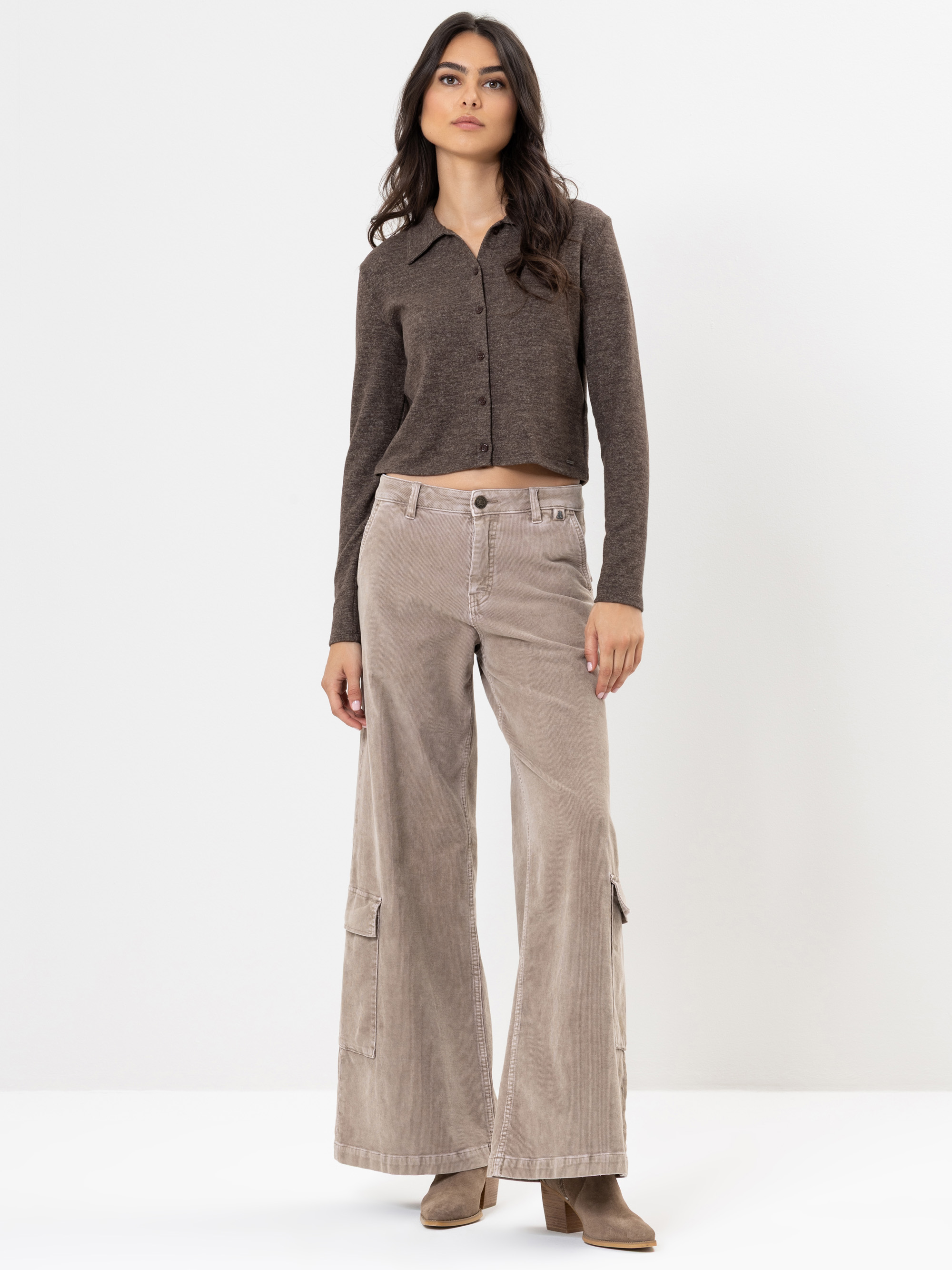 Eine Frau steht vor einem weißen Hintergrund und trägt ein braunes, langärmeliges, geknöpftes Oberteil, eine beigefarbene Cargohose mit weitem Bein und taupefarbene Ankle Boots. Sie hat langes, dunkles Haar und einen neutralen Gesichtsausdruck.