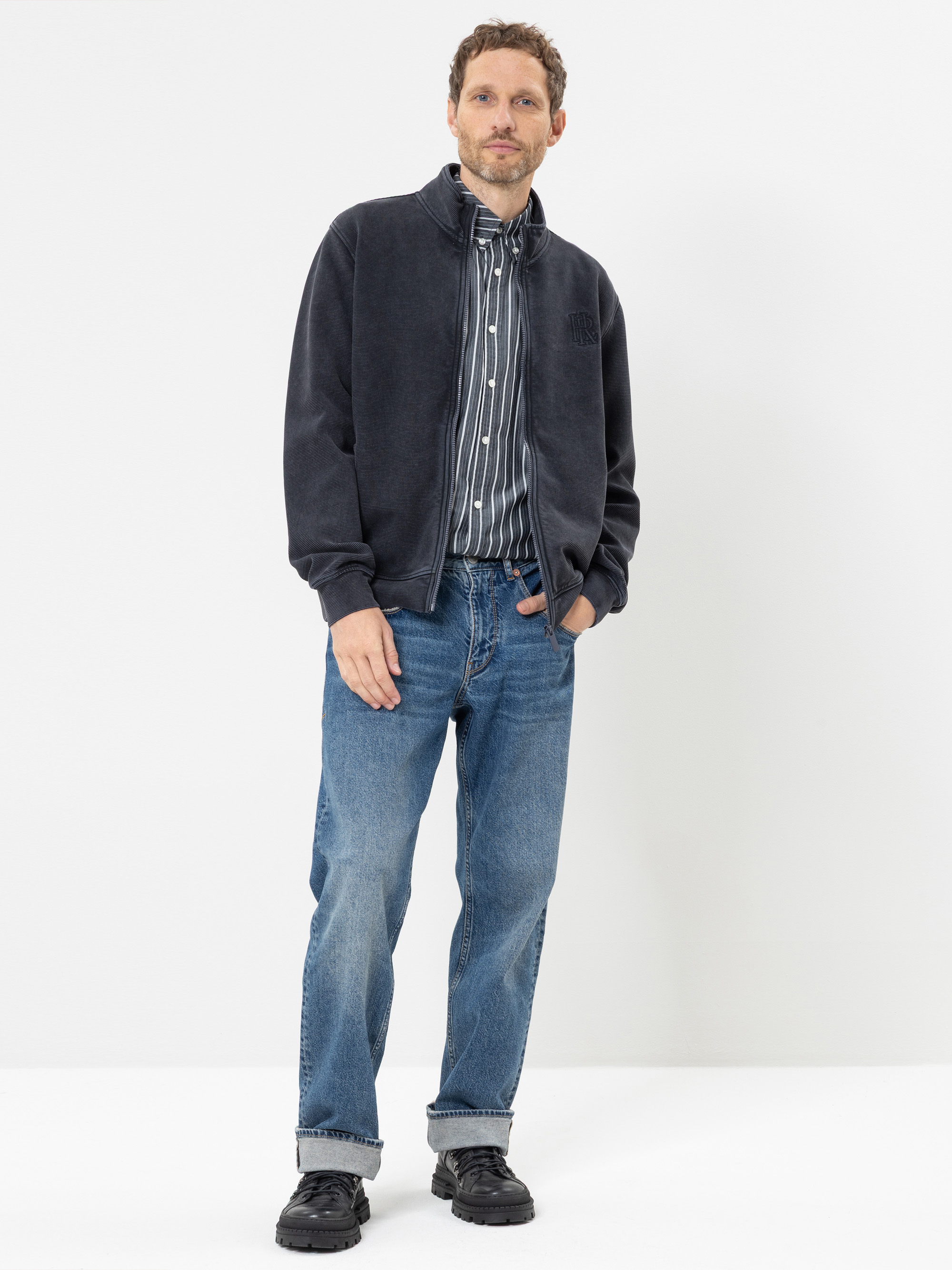 Vor einem weißen Hintergrund steht ein Mann, der eine dunkle Jacke mit Reißverschluss, ein gestreiftes Hemd mit Knöpfen, blaue Jeans und schwarze Schuhe trägt. Er hat eine Hand in der Tasche und blickt direkt in die Kamera.