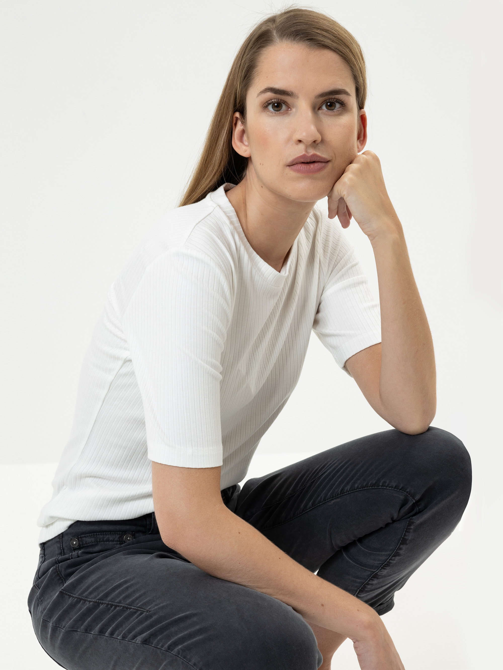 Eine Person mit langem, glattem braunem Haar sitzt in einer entspannten Pose und stützt ihr Kinn auf eine Hand. Sie trägt ein weißes, kurzärmeliges Oberteil und eine dunkle Jeans und blickt selbstbewusst in die Kamera vor einem schlichten Hintergrund.