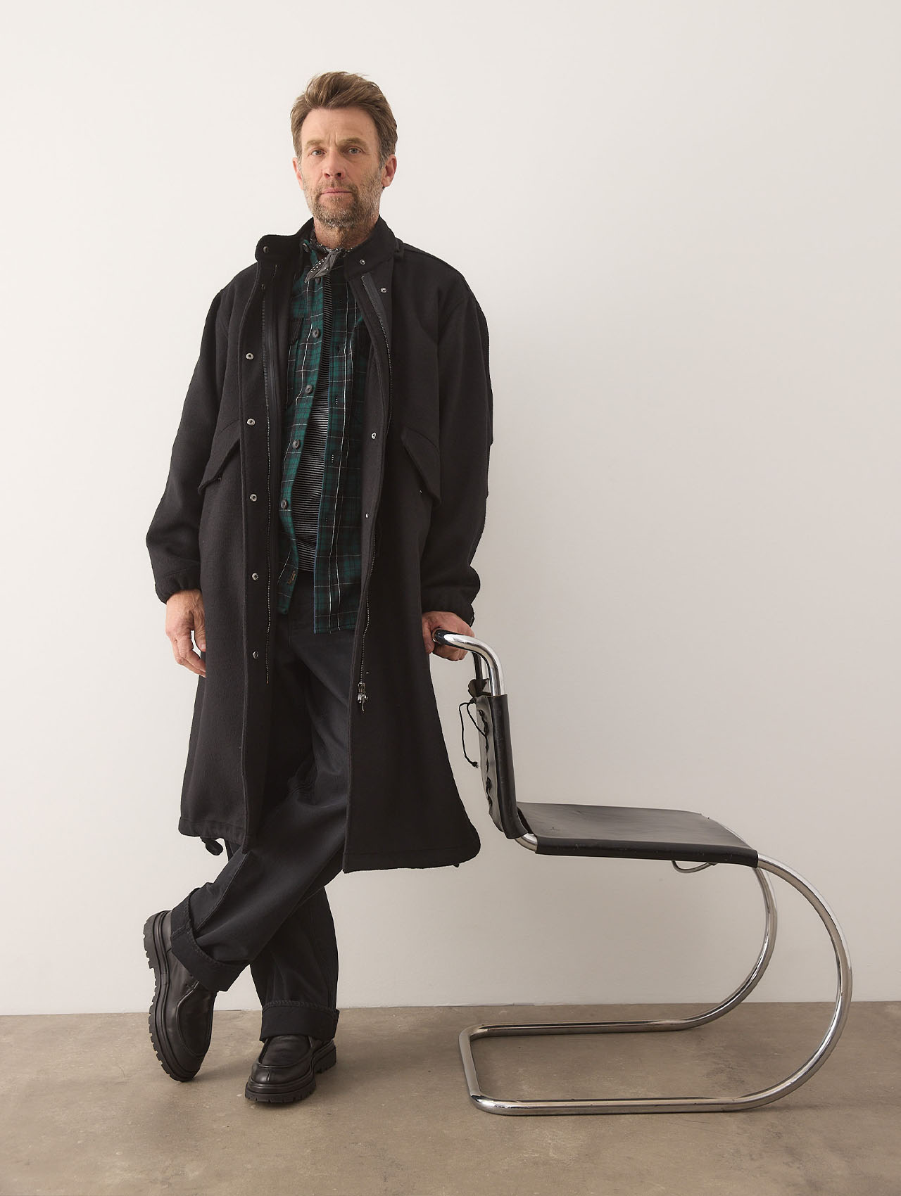 Ein Mann, der einen langen schwarzen Mantel, ein grün kariertes Hemd, eine schwarze Hose und schwarze Schuhe trägt, steht neben einem modernen schwarz-verchromten Stuhl vor einer schlichten weißen Wand.