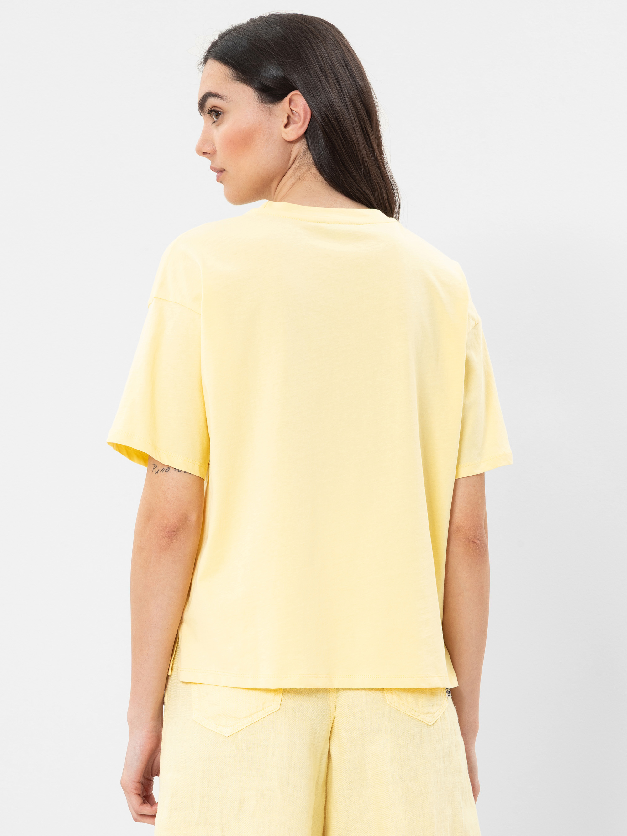 Eine Frau mit langen dunklen Haaren steht mit dem Gesicht zur Wand und trägt ein locker sitzendes gelbes T-Shirt und passende gelbe Shorts vor einem schlichten weißen Hintergrund.