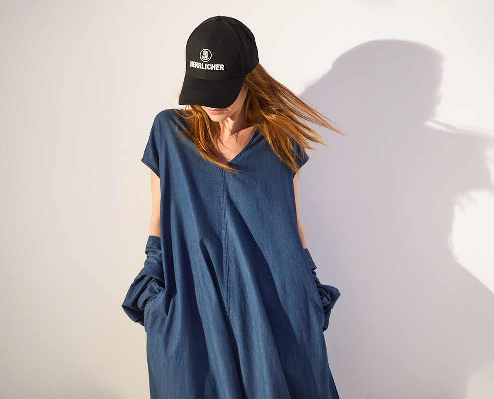 Eine Person mit langen Haaren trägt ein locker sitzendes blaues Kleid und eine schwarze Mütze mit weißer Aufschrift und steht vor einer weißen Wand mit Schatten. Ihr Kopf ist nach unten geneigt, wodurch ihr Gesicht teilweise verdeckt wird.