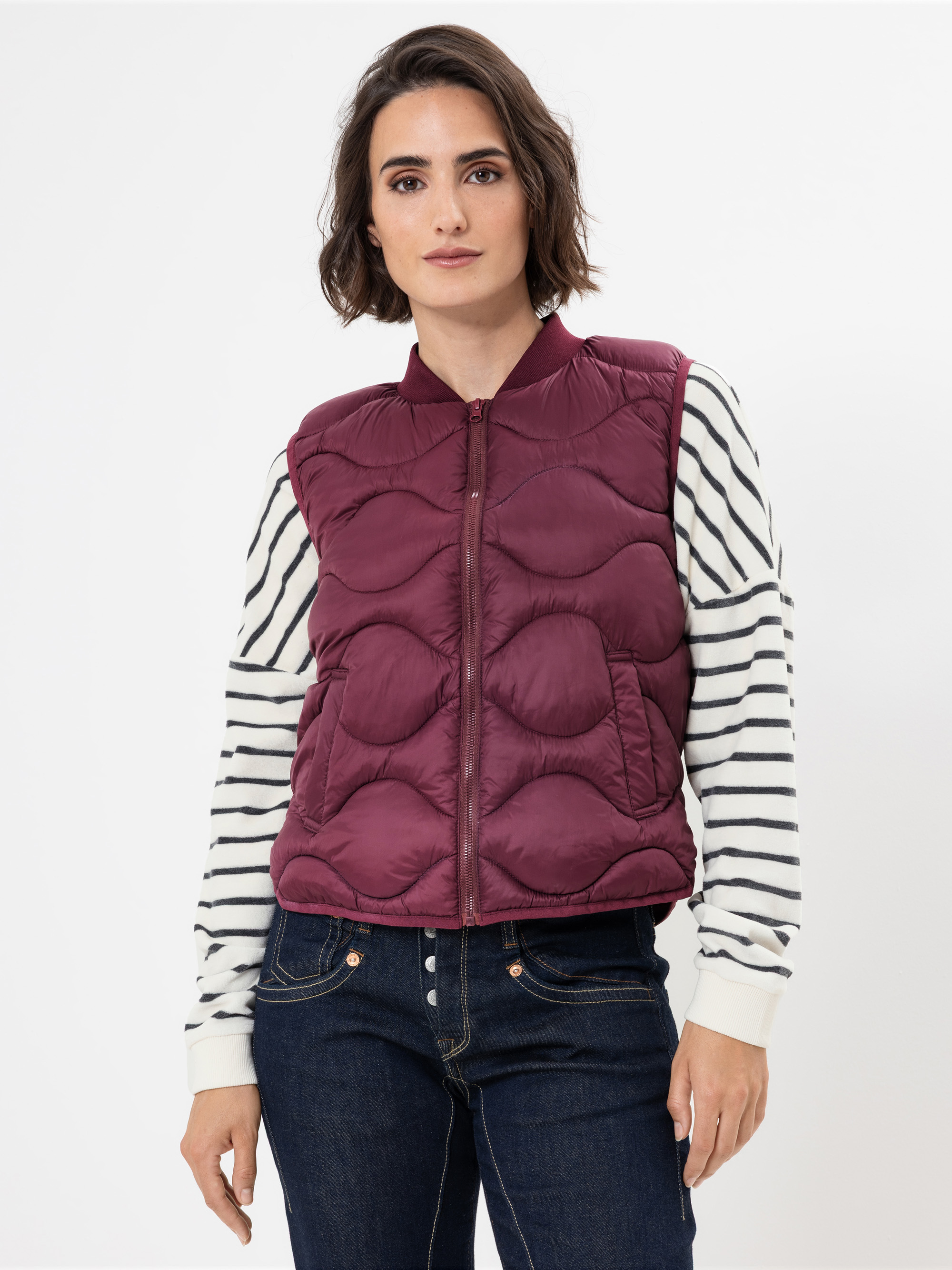 Eine Frau mit kurzen braunen Haaren trägt eine kastanienbraune Steppweste über einem weißen langärmeligen Hemd mit schwarzen Querstreifen, dazu dunkelblaue Jeans, vor einem schlichten weißen Hintergrund.