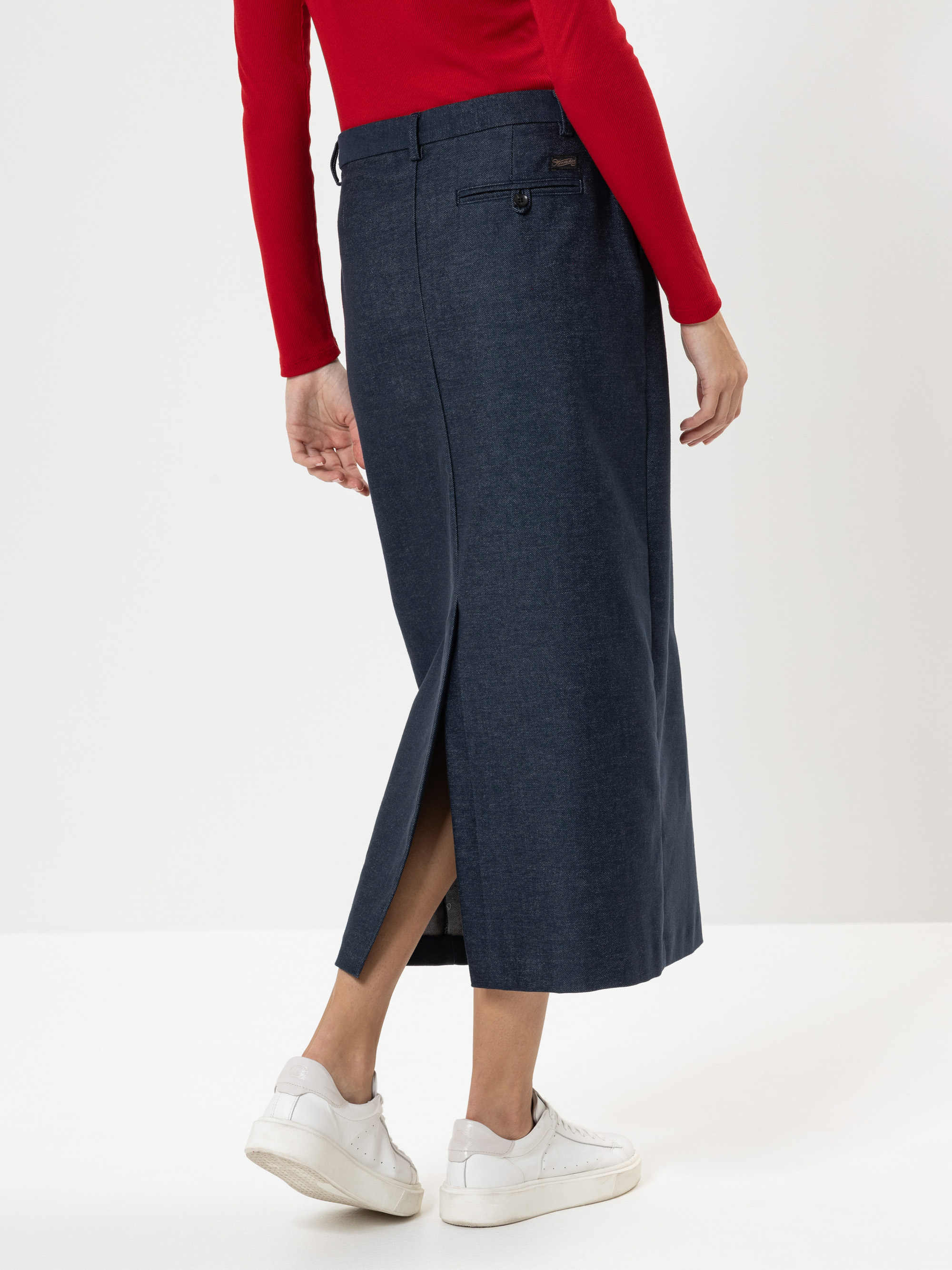 Eine Person, die ein rotes langärmeliges Oberteil, einen dunkelblauen Jeans-Midirock mit Rückenschlitz und weiße Turnschuhe trägt, steht vor einem schlichten weißen Hintergrund.