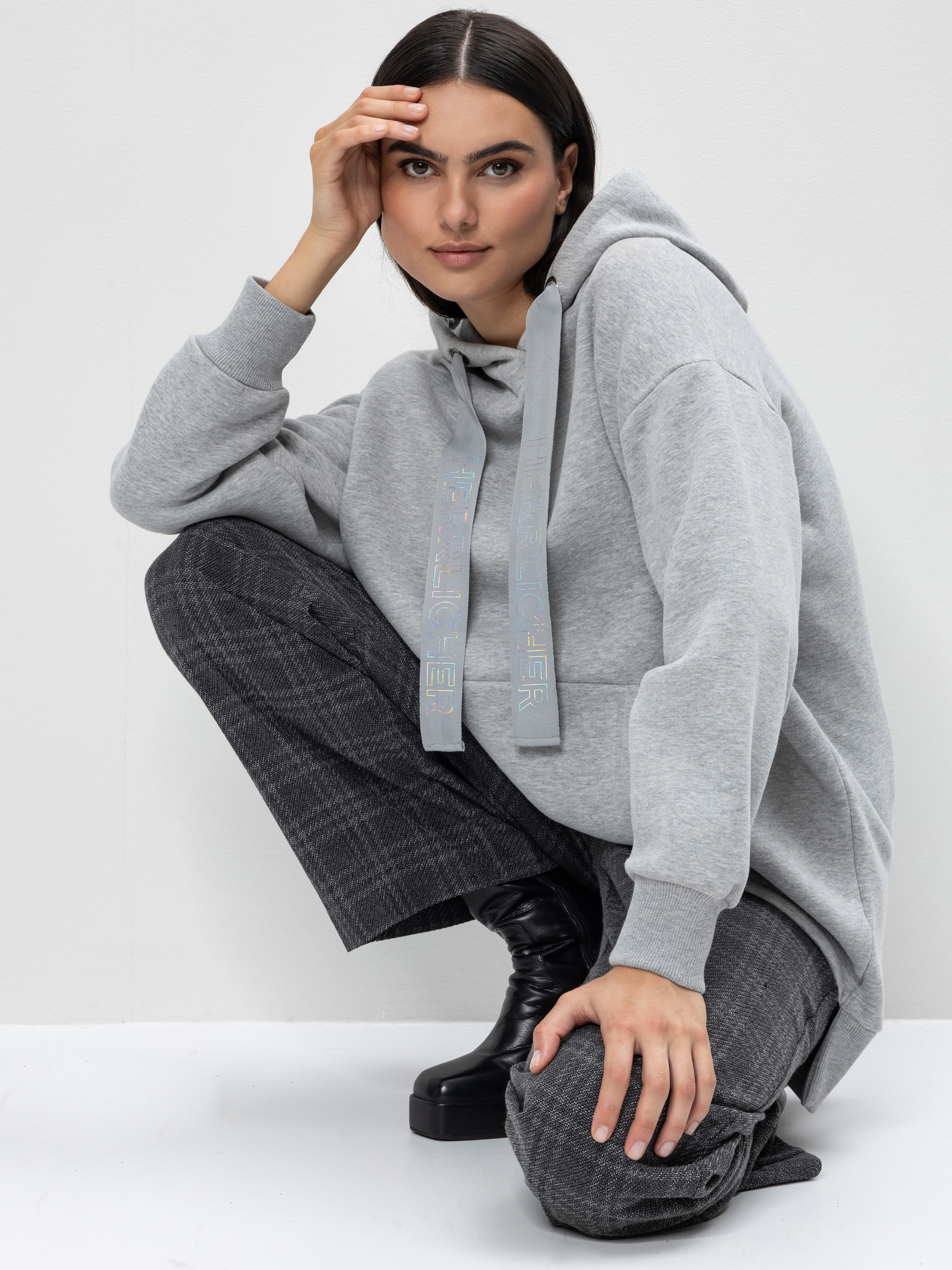 Eine Frau mit langen dunklen Haaren, die einen grauen Kapuzenpulli, eine dunkelkarierte Hose und schwarze Stiefel trägt, geht in die Hocke und stützt eine Hand auf ihr Knie, während die andere ihre Stirn berührt.