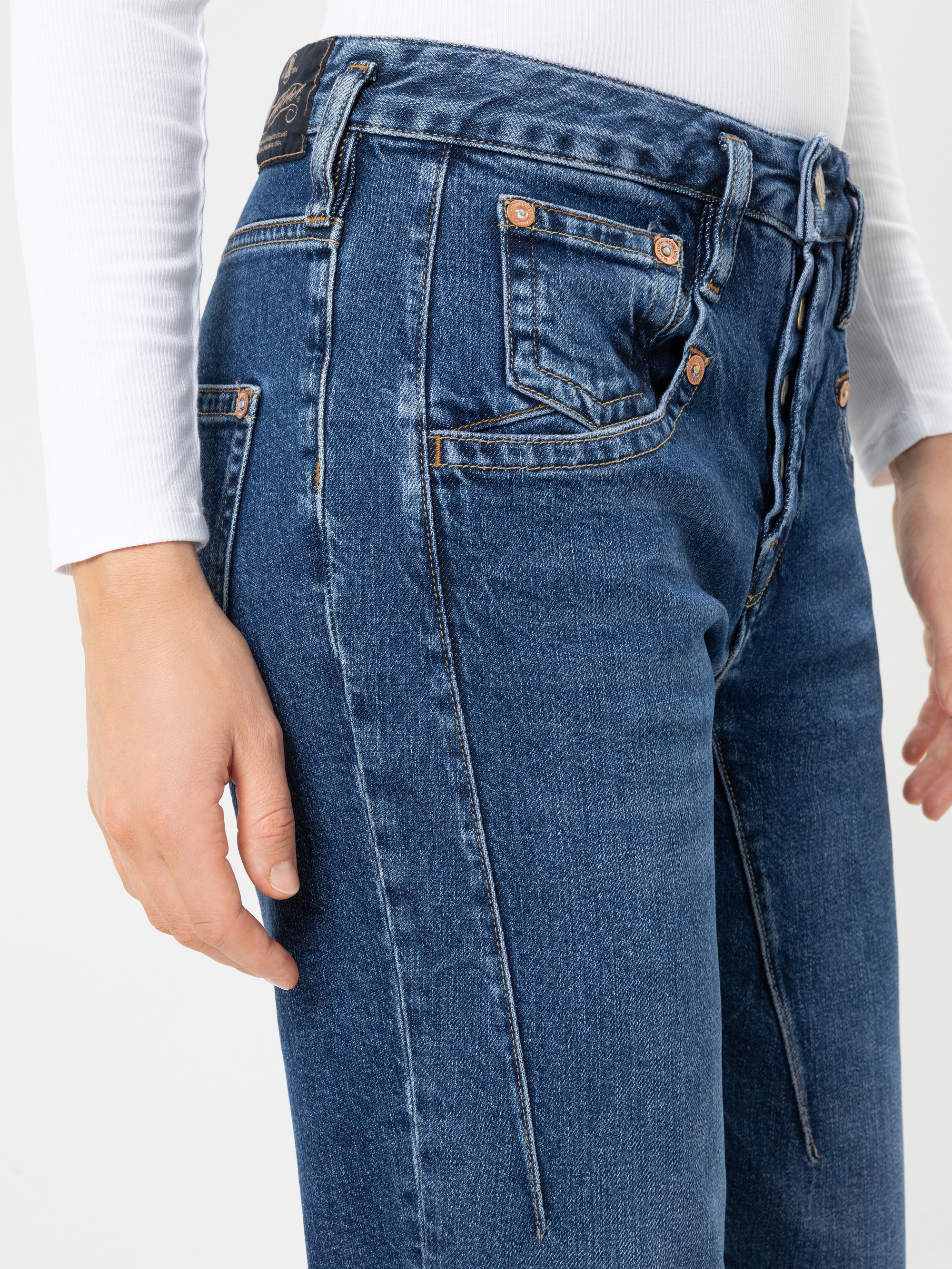 Eine Person in einem weißen Langarm-Oberteil und einer hoch taillierten blauen Jeans mit sichtbaren Vordernähten und kupferfarbenen Nieten, die vor einem einfarbigen Hintergrund steht.