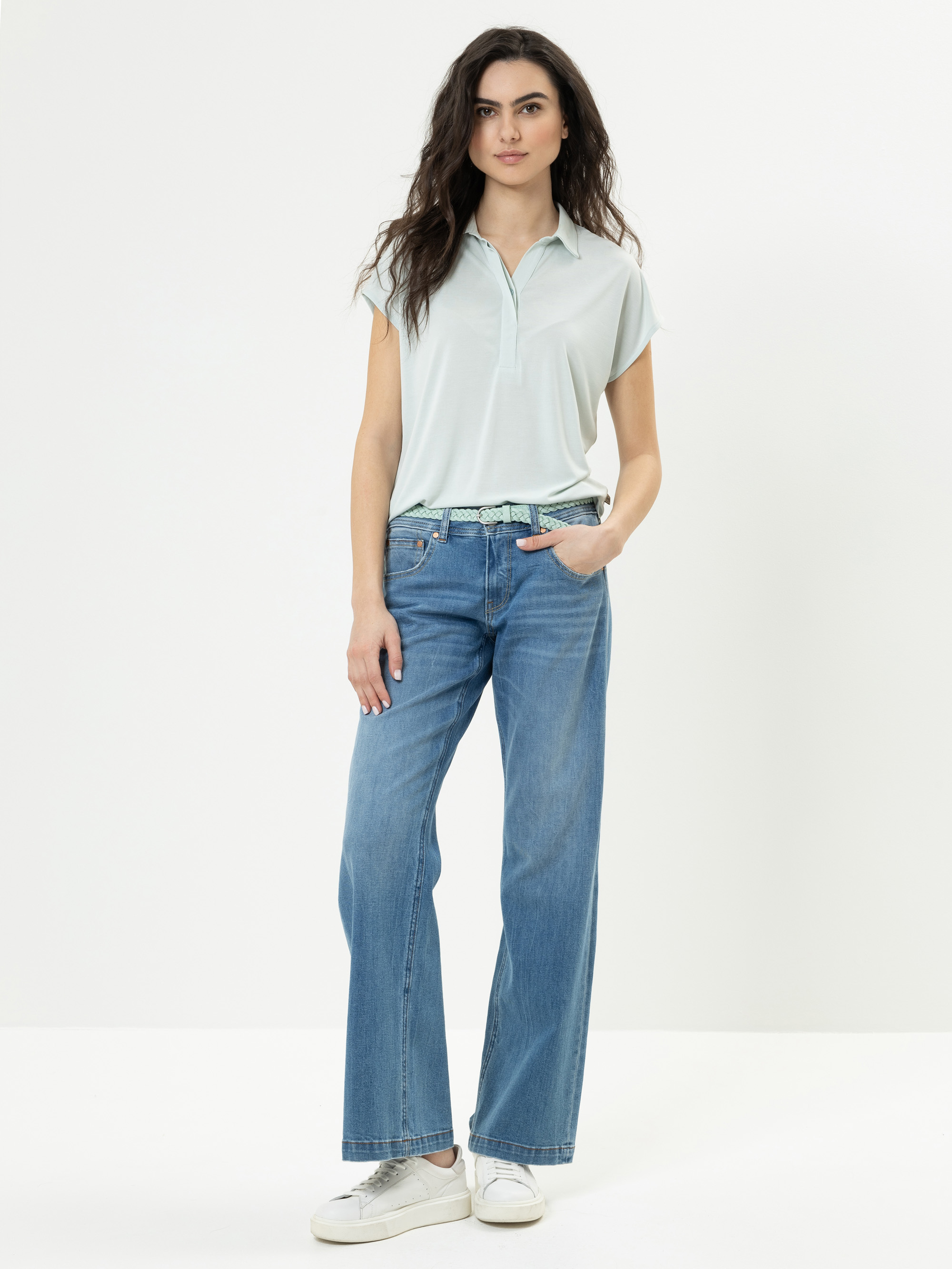 Eine Frau mit langen dunklen Haaren steht vor einem einfarbig weißen Hintergrund. Sie trägt ein hellblaues, kurzärmeliges Polohemd, eine blaue Jeans mit weitem Bein, einen gemusterten Gürtel und weiße Turnschuhe, eine Hand steckt in ihrer Tasche.
