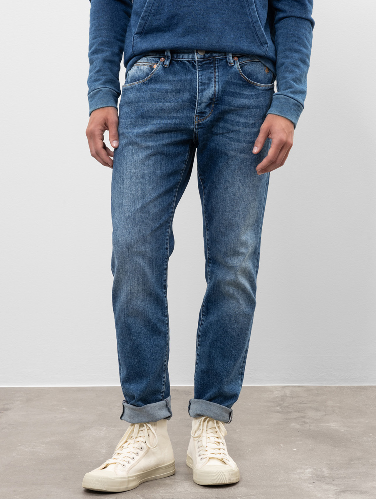 Eine Person, die ein blaues Sweatshirt, eine blaue Jeans mit aufgerollten Manschetten und weiße High-Top-Turnschuhe trägt, steht auf einem Betonboden vor einer schlichten weißen Wand.