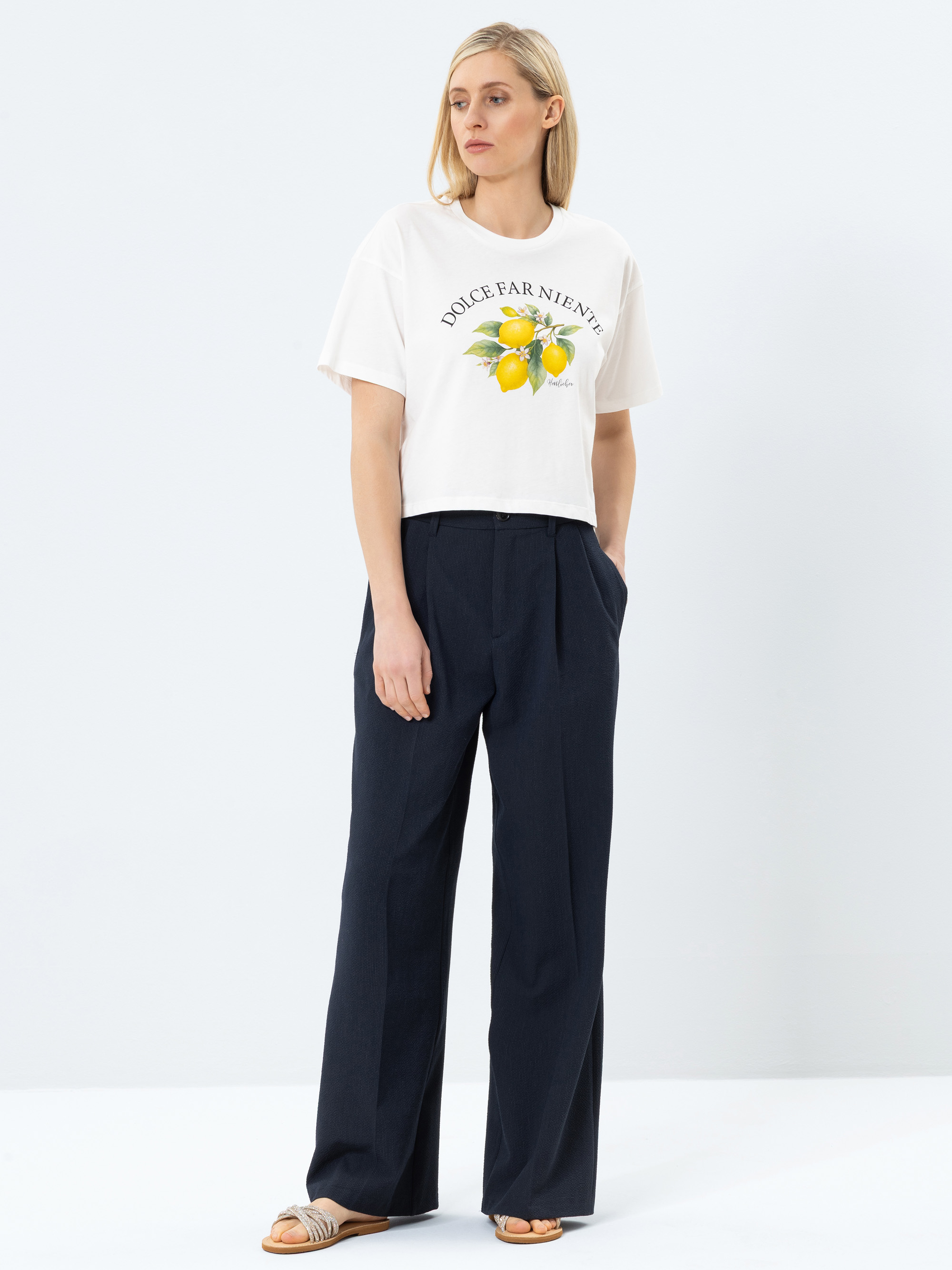 Eine Frau mit blondem Haar trägt ein weißes T-Shirt von Dolce Far Niente mit einer Zitronengrafik, eine marineblaue Hose mit weitem Bein und Sandalen. Sie steht mit den Händen in den Taschen vor einem schlichten weißen Hintergrund.