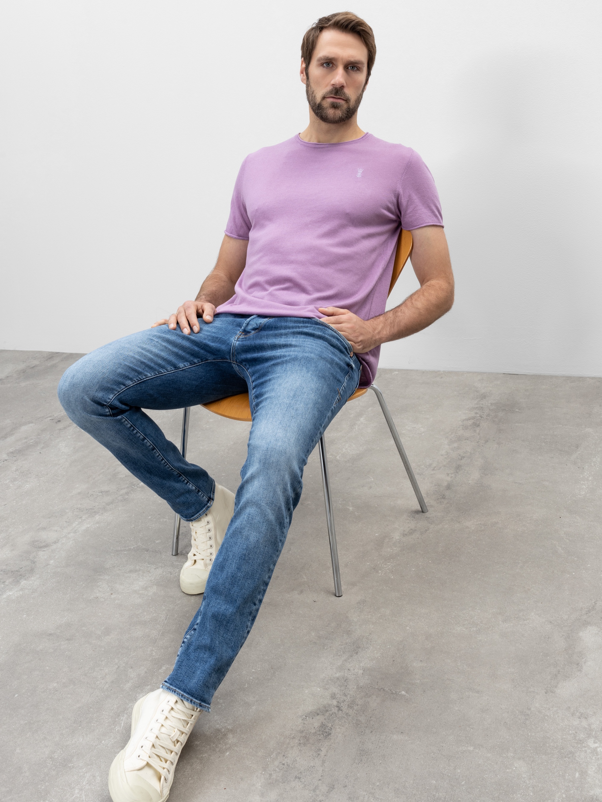 Ein Mann mit kurzen Haaren und einem Bart sitzt auf einem Holzstuhl und trägt ein lavendelfarbenes T-Shirt, blaue Jeans und weiße Turnschuhe. Der Raum hat einen schlichten grauen Boden und weiße Wände.