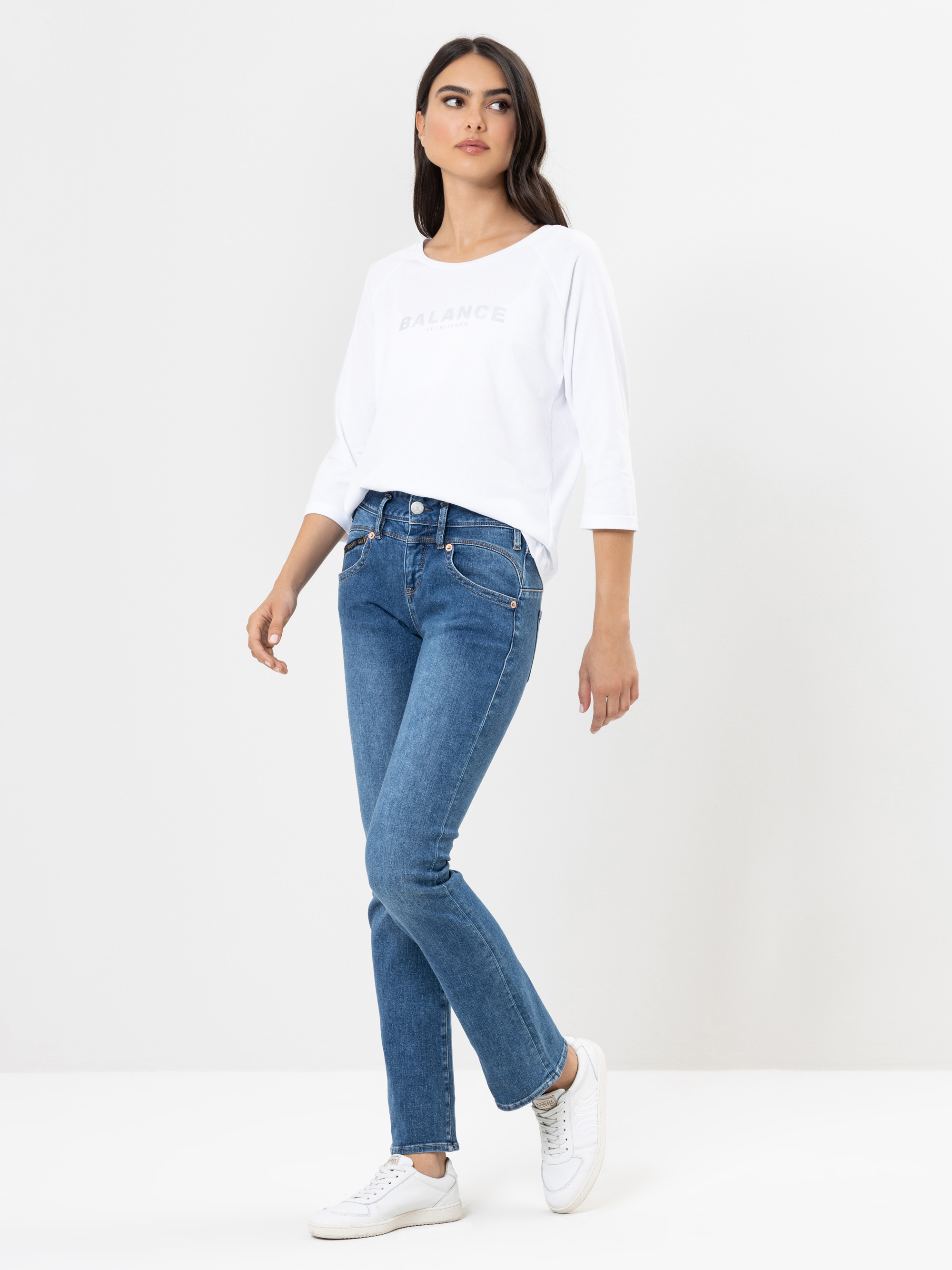 Eine Frau steht vor einem schlichten weißen Hintergrund und trägt ein weißes, langärmeliges Hemd mit der Aufschrift BALANCE, blaue Jeans und weiße Turnschuhe. Sie hat lange dunkle Haare und posiert mit einem leicht angewinkelten Bein.
