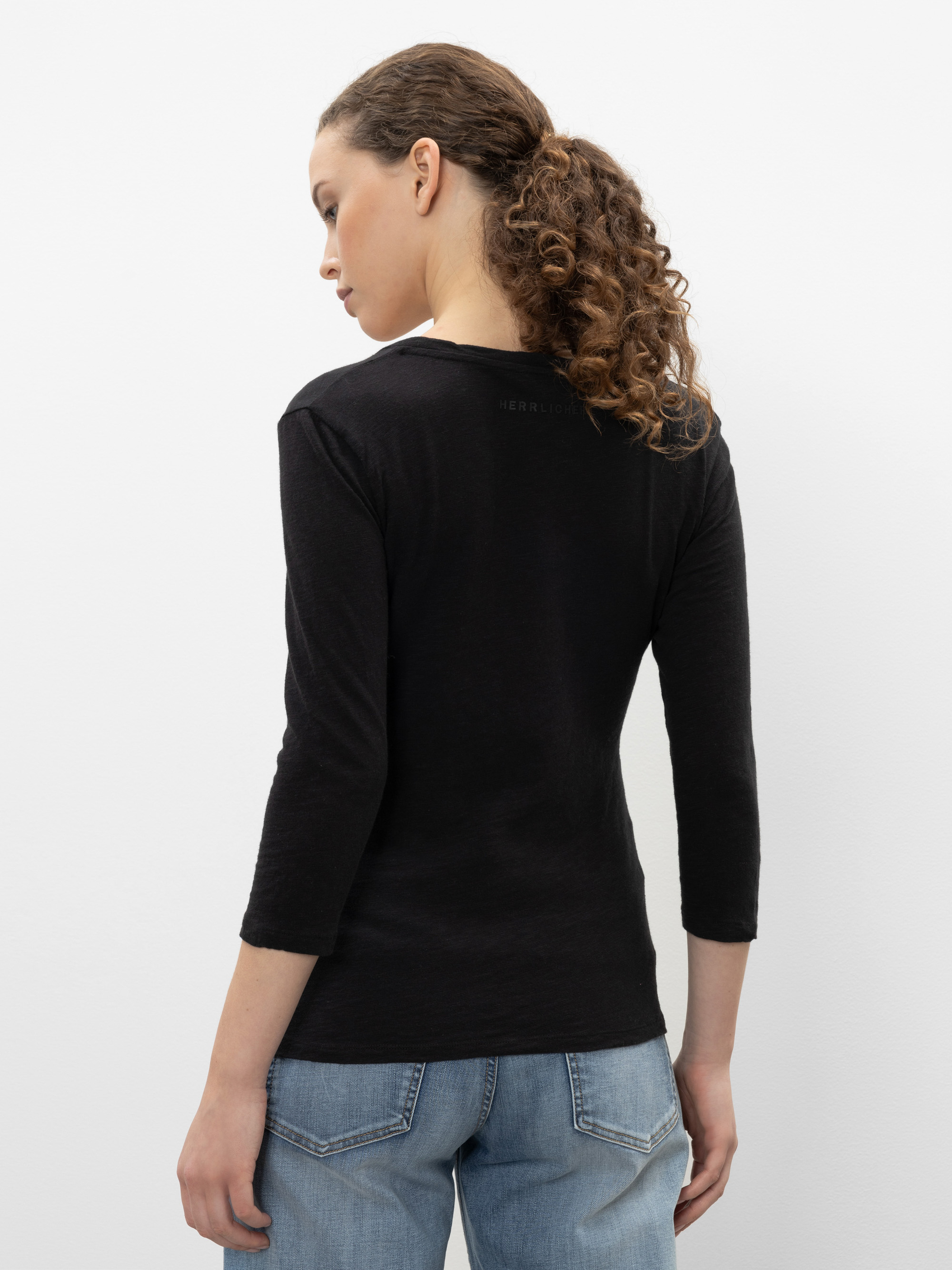 Eine Frau mit lockigem Haar in einem Pferdeschwanz steht mit dem Rücken zur Kamera, trägt ein schwarzes dreiviertelärmliges Oberteil und hellblaue Jeans vor einem schlichten weißen Hintergrund.