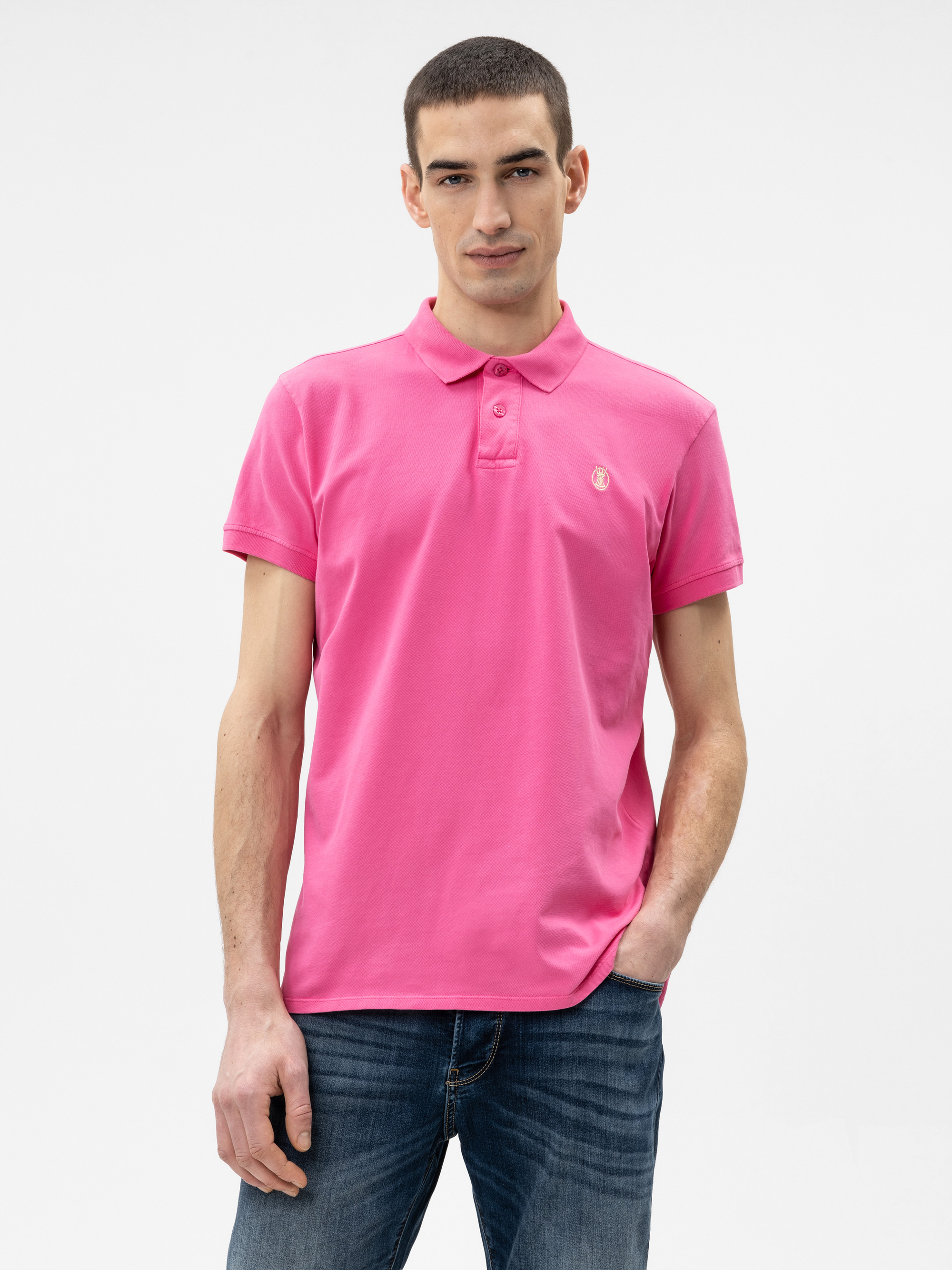 Ein Mann in einem leuchtend rosafarbenen Polohemd und einer blauen Jeans steht vor einem schlichten weißen Hintergrund, eine Hand in der Tasche und mit einem neutralen Gesichtsausdruck.