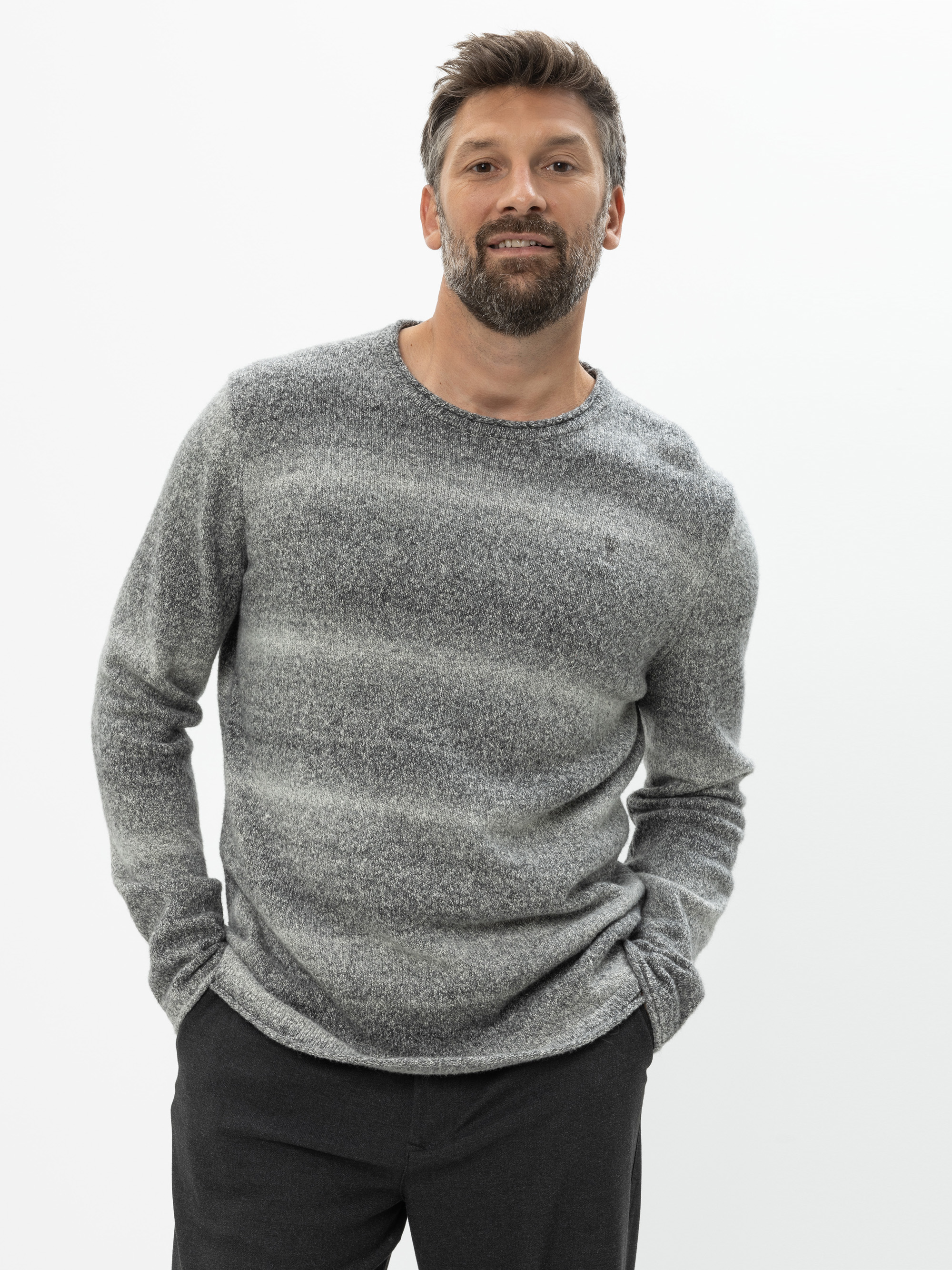 Ein Mann mit kurzen braunen Haaren und einem Bart trägt einen grauen, langärmeligen Pullover und eine dunkle Hose. Er steht vor einem schlichten weißen Hintergrund, hat die Hände in den Taschen, blickt leicht nach oben und lächelt.