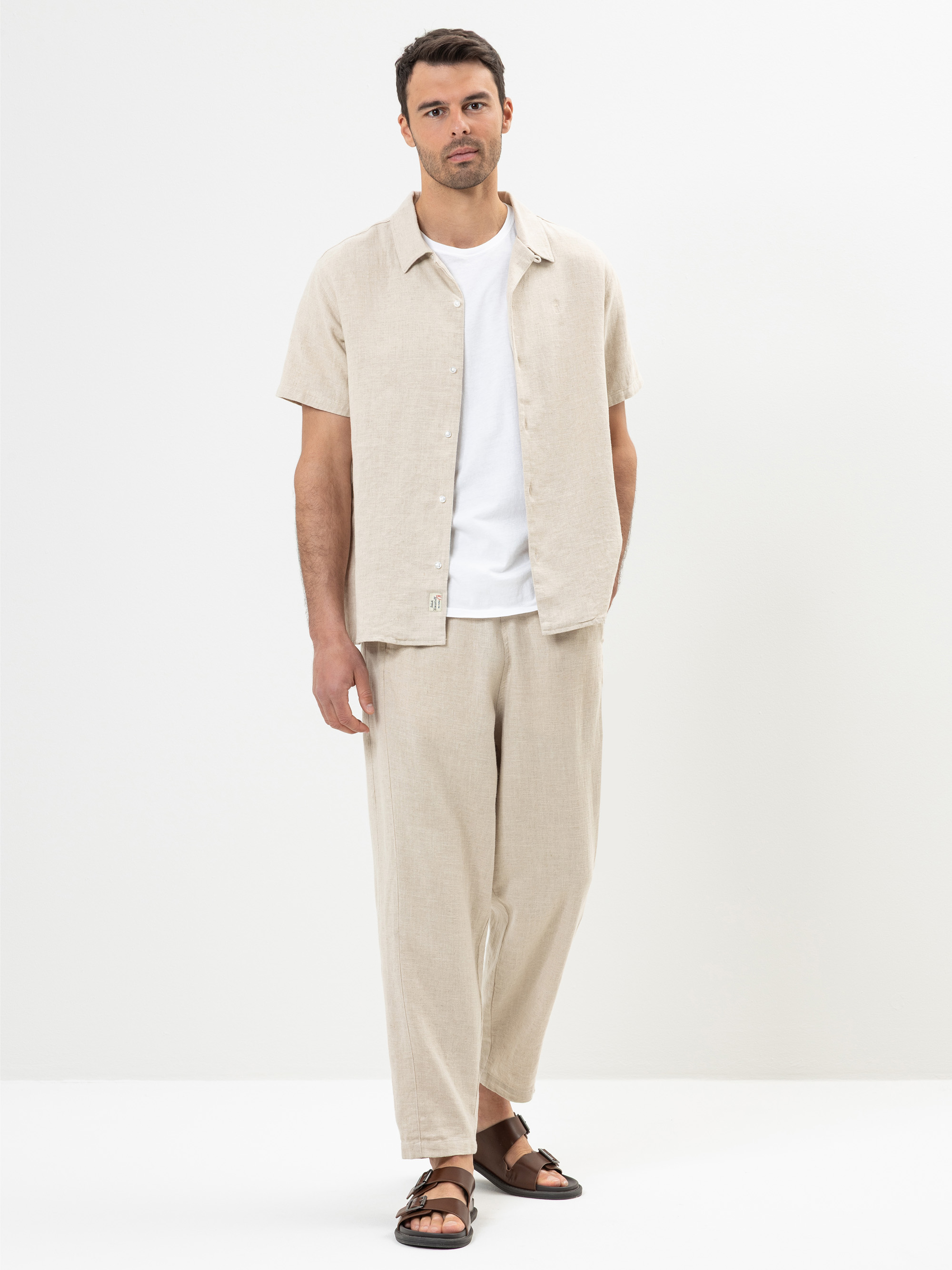 Ein Mann steht vor einem schlichten Hintergrund und trägt ein beigefarbenes, kurzärmeliges Button-up-Hemd, eine dazu passende weite Hose, darunter ein weißes T-Shirt und braune Sandalen. Eine Hand steckt in seiner Tasche und er schaut in die Kamera.